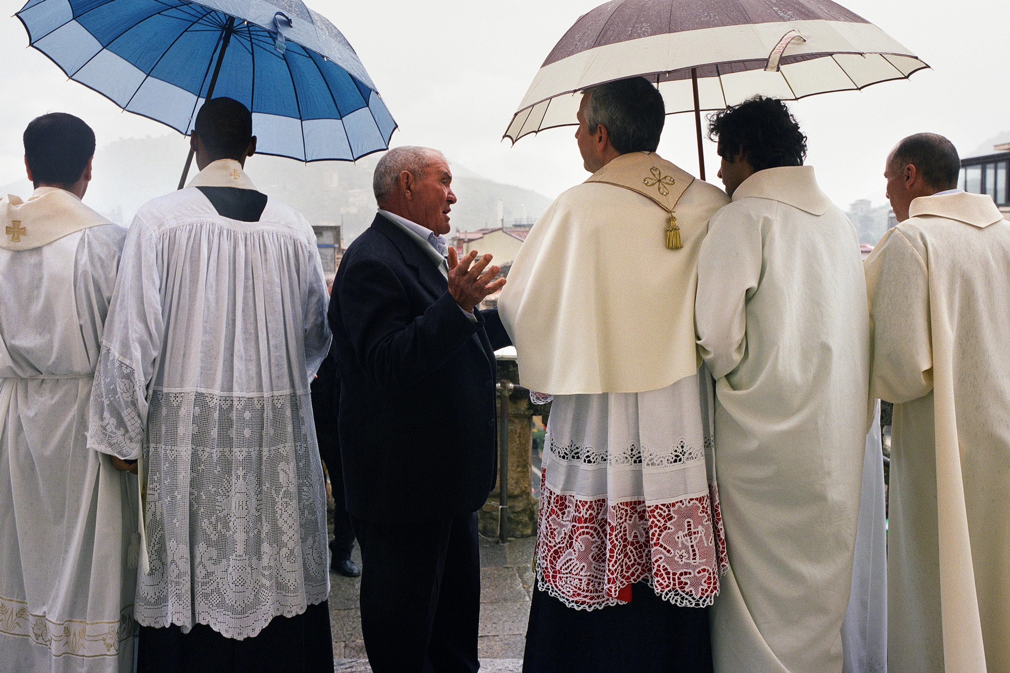 Photo of clergy in white robes and a man with umbrellas, standing outdoors on a rainy day.