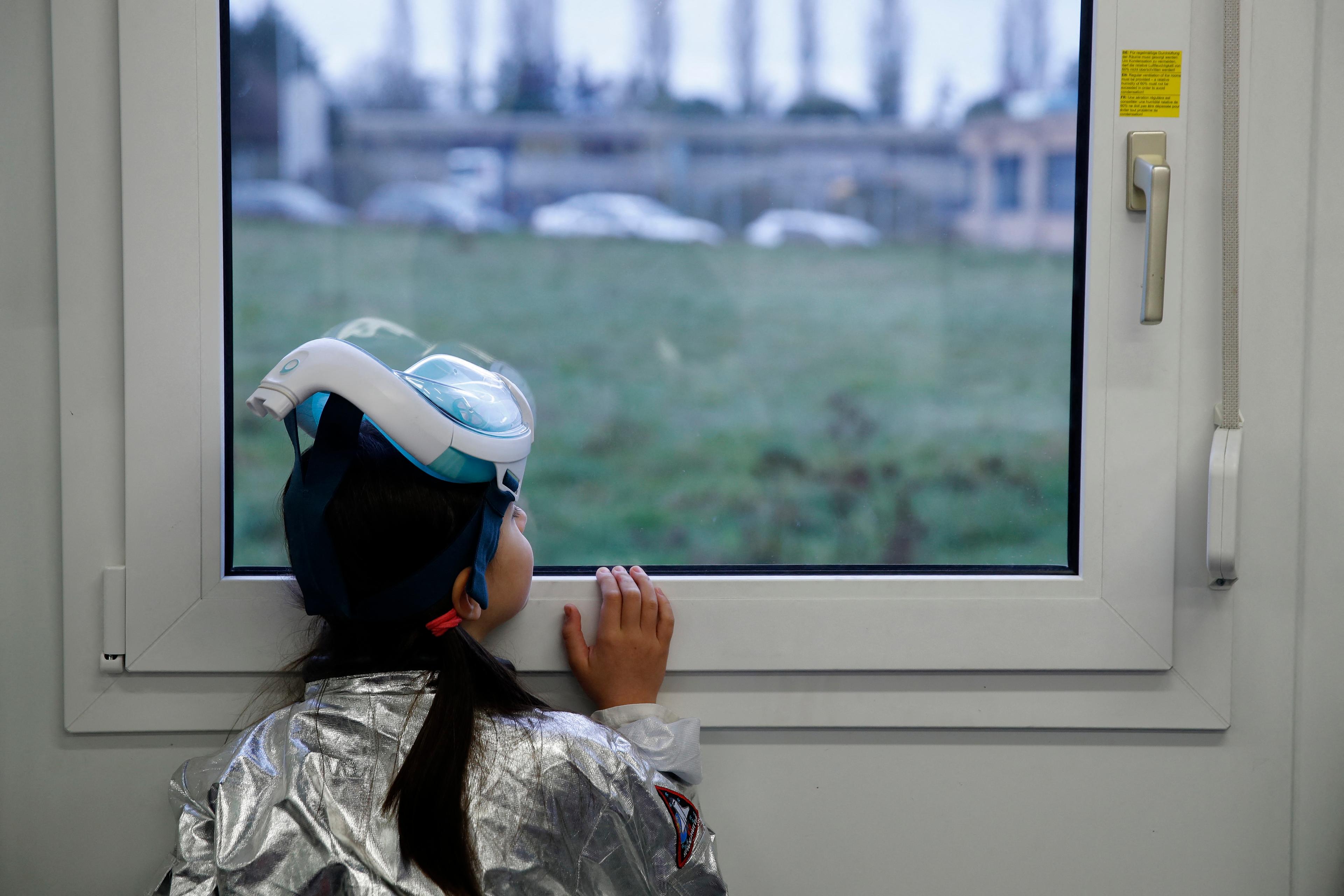 Photo of a child in a silver suit and snorkelling mask looking out of a window at a grassy landscape.