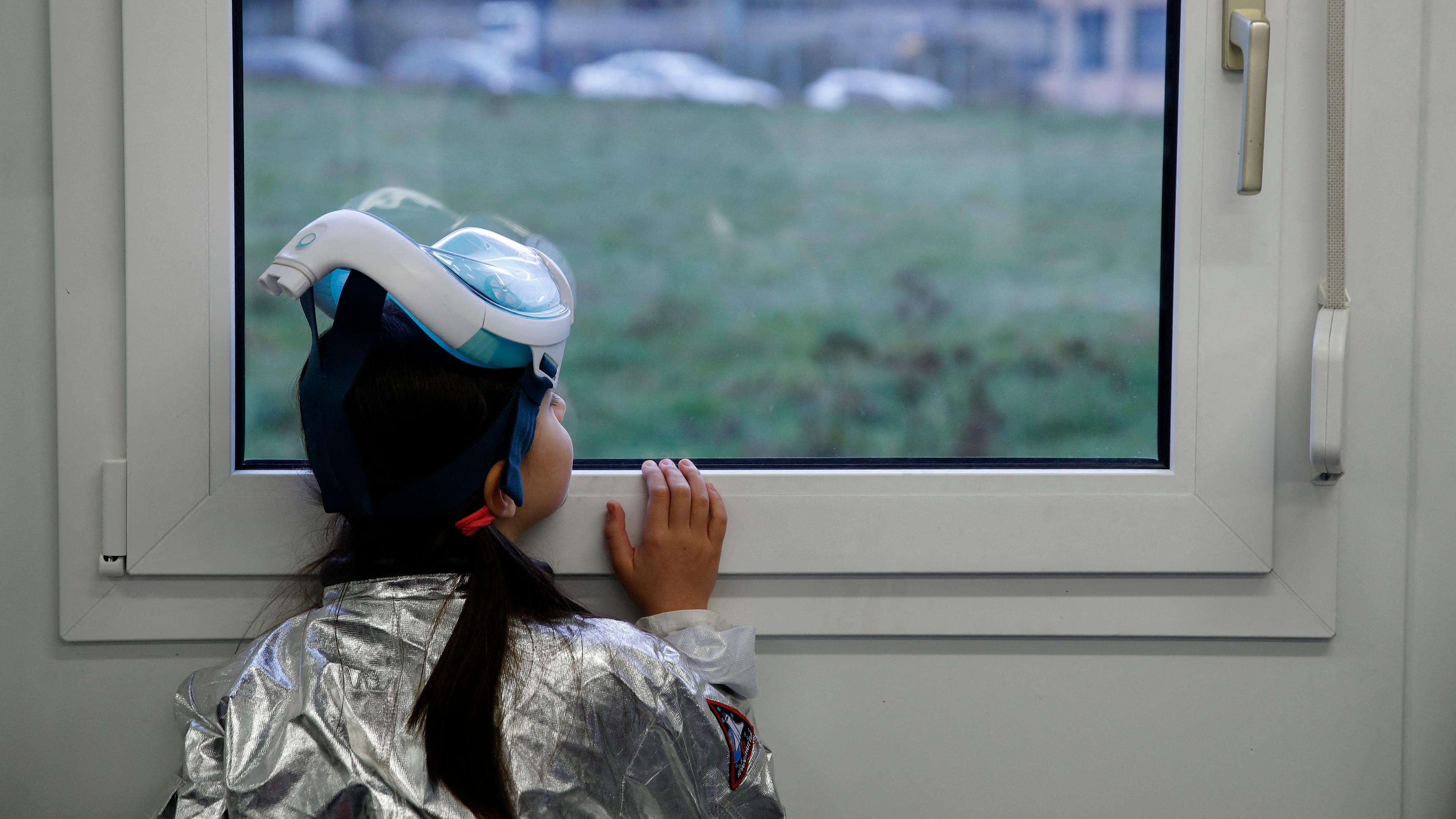Photo of a child in a silver suit and snorkelling mask looking out of a window at a grassy landscape.