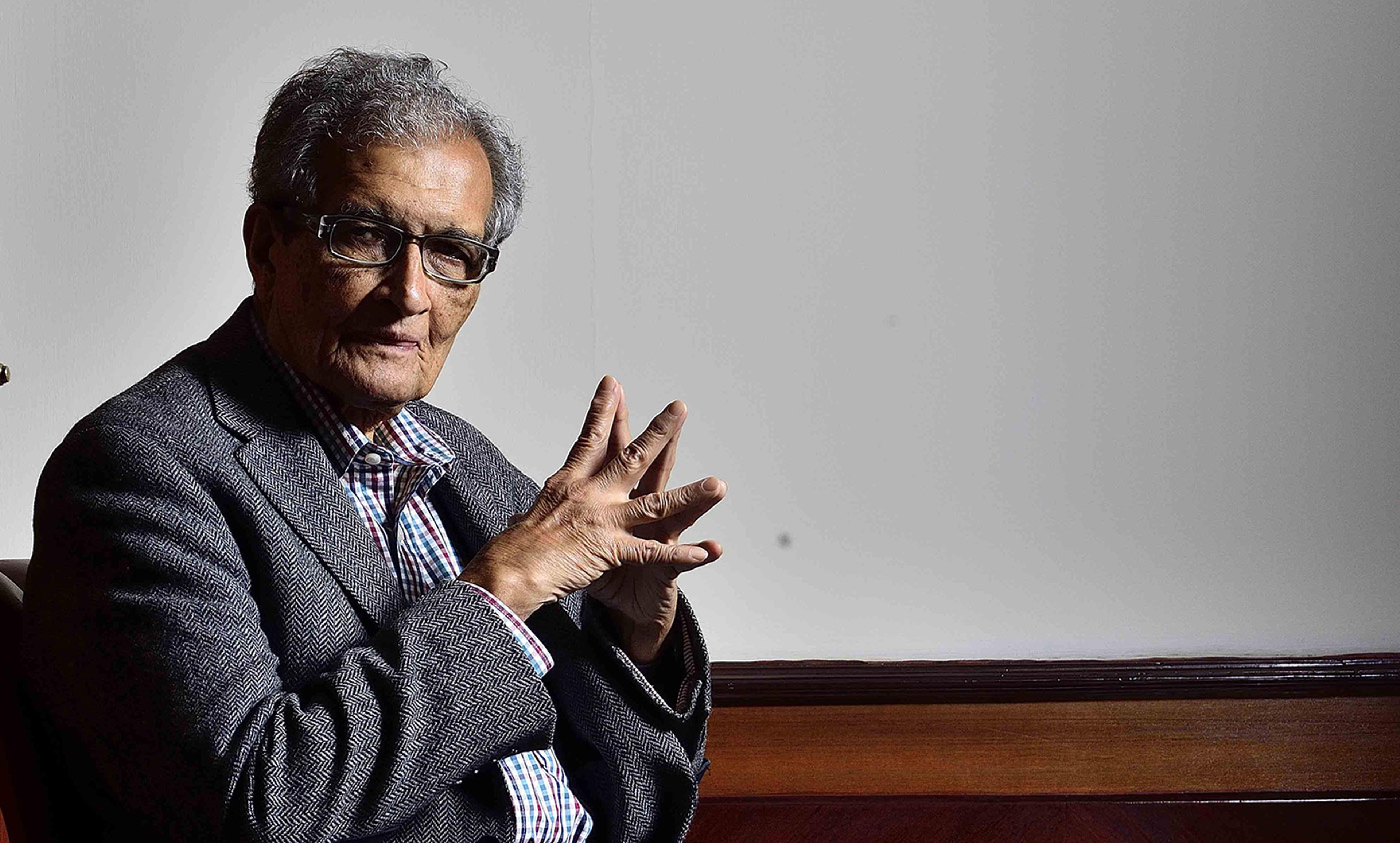 Amartya Sen with glasses and grey hair wearing a checked shirt and grey blazer, sitting with his hands clasped.