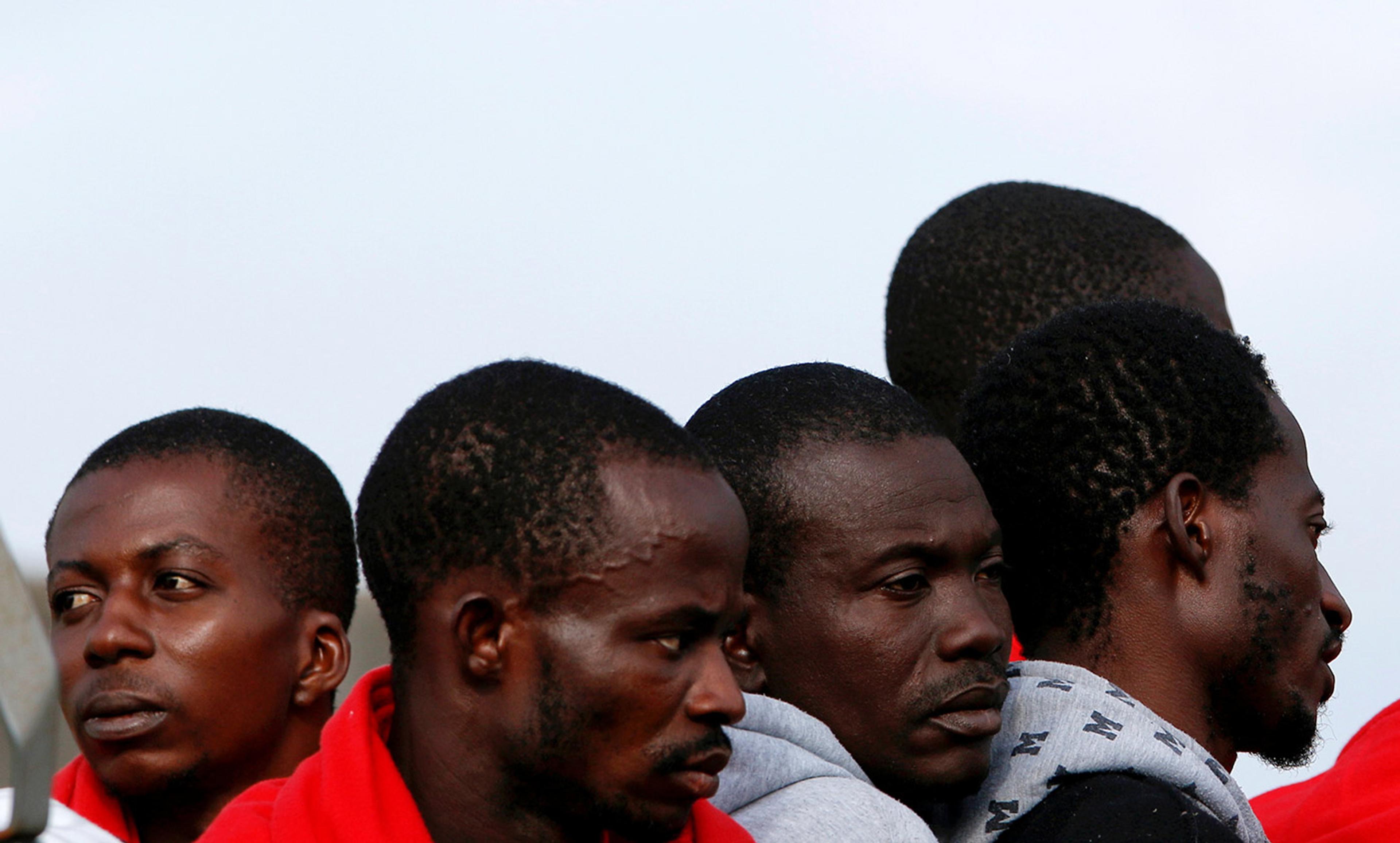 Five men standing closely together, some wearing hoodies and red jackets, looking in different directions. A clear sky background.