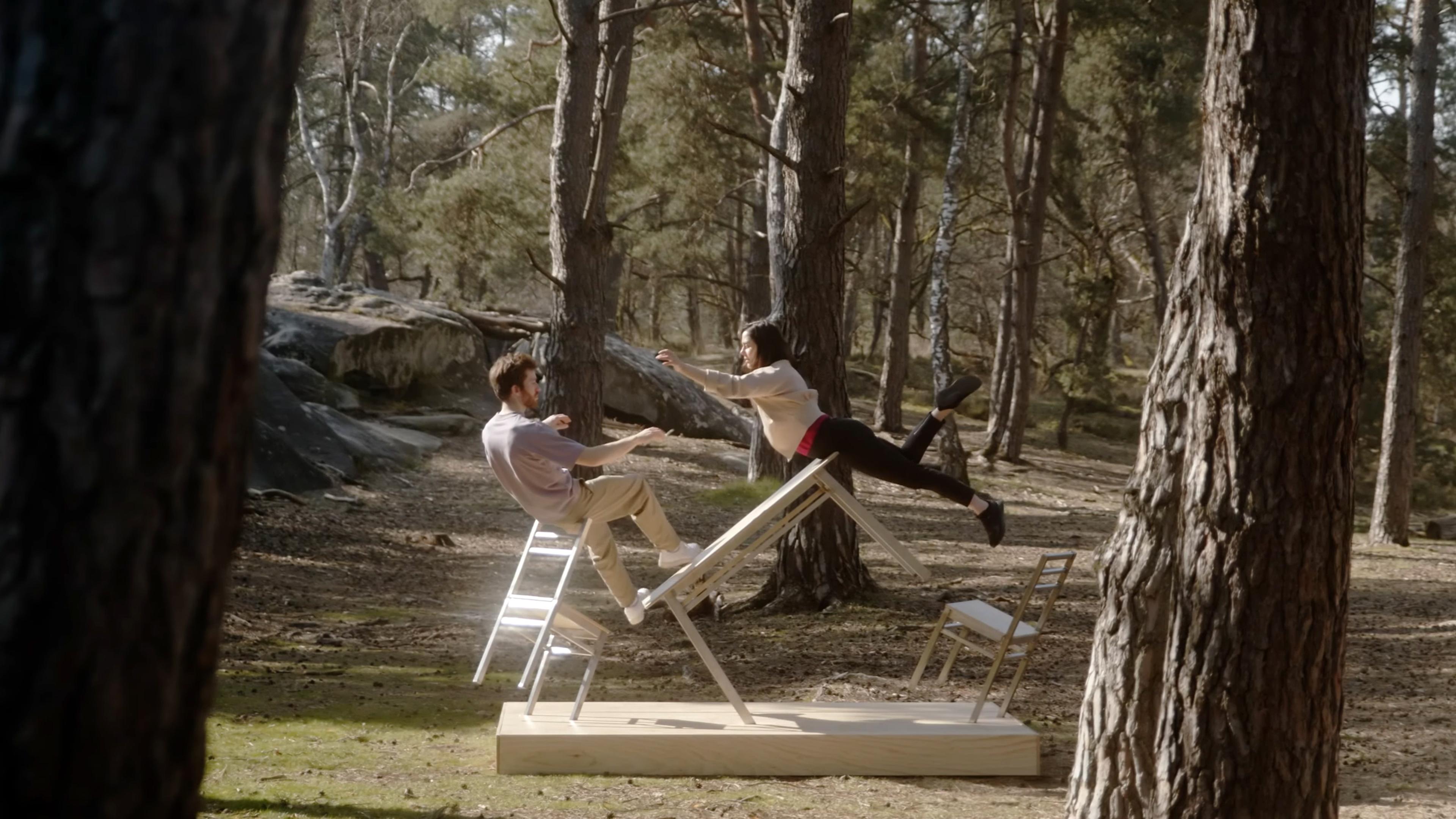 Two people in a park performing a balancing act on white chairs and a wooden plank, surrounded by pine trees.