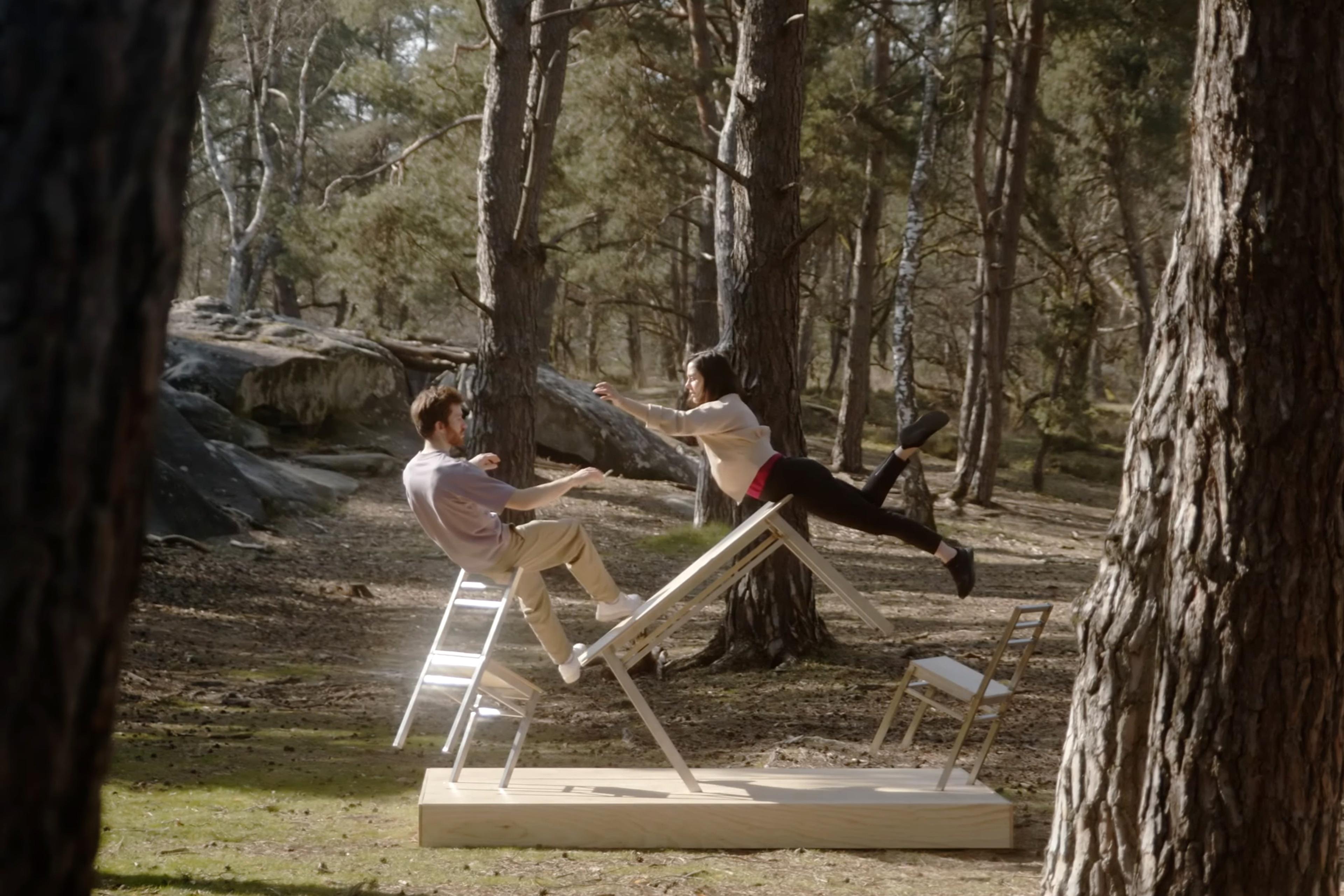 Two people in a park performing a balancing act on white chairs and a wooden plank, surrounded by pine trees.