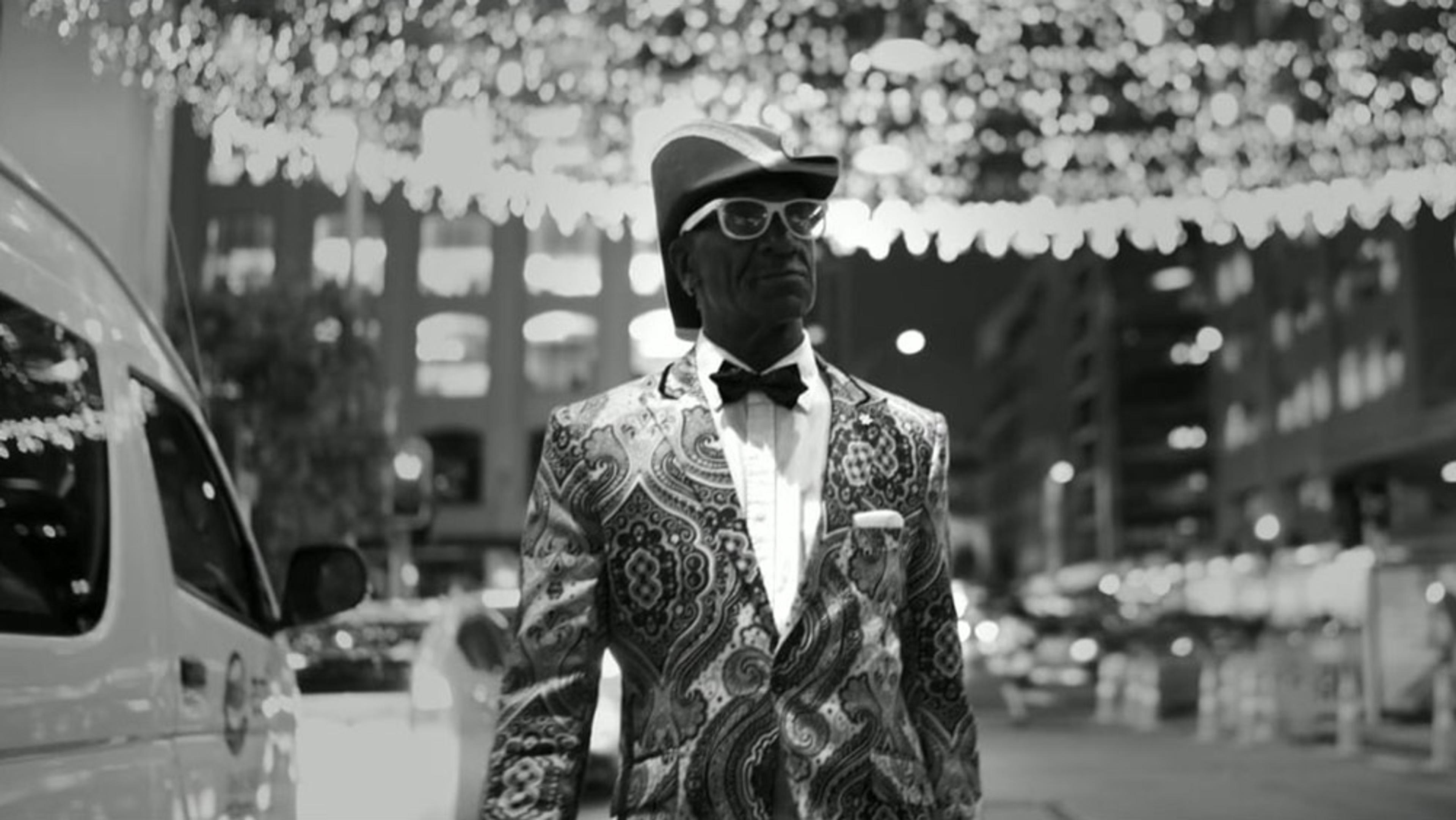 Black-and-white photo of a man in a patterned suit, bow tie and hat, standing on a lit street at night with buildings in the background.