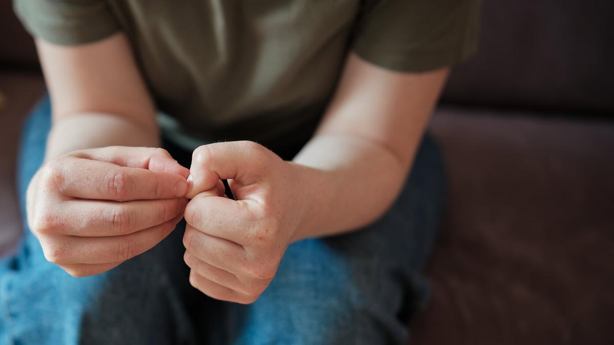 A person in a green shirt and jeans sitting, focused on their hands which are clasped together, the right hand picking at the left hand’s thumbnail.