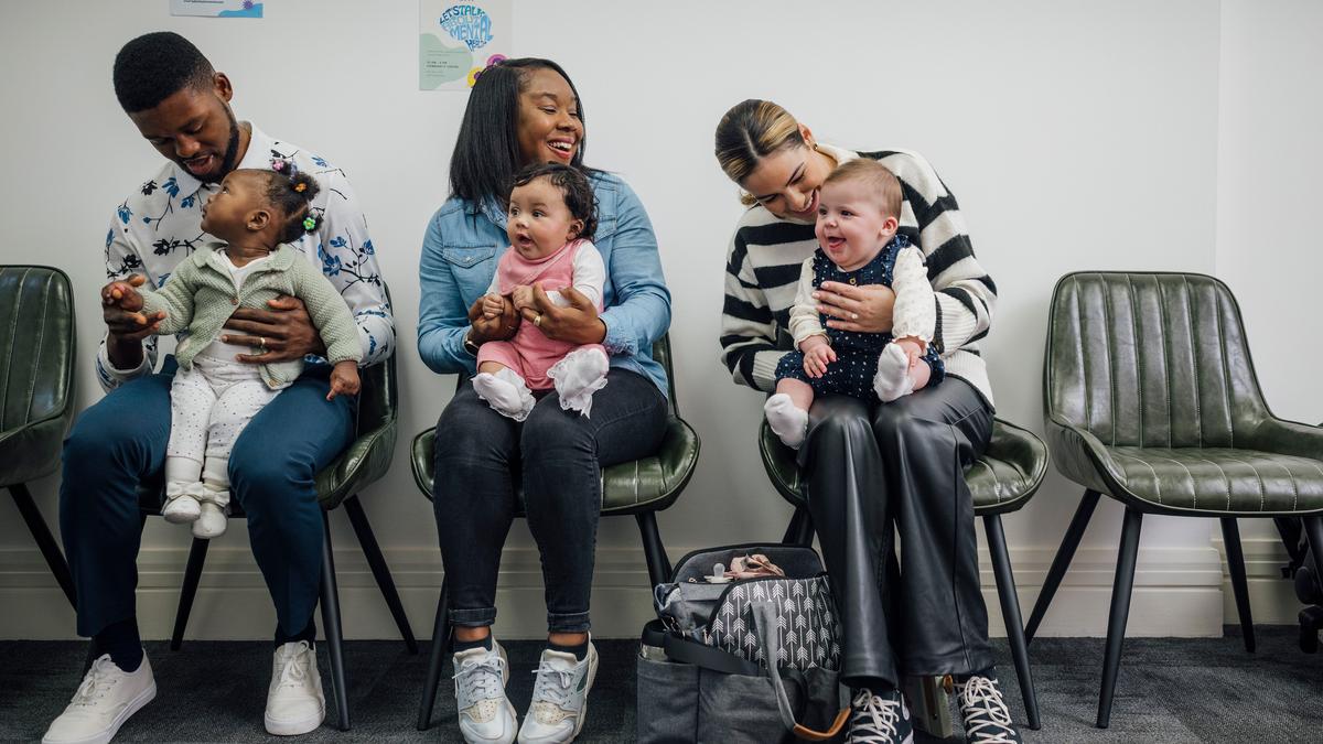 Three adults with babies seated in a waiting room, smiling and interacting, with colourful posters on the wall.