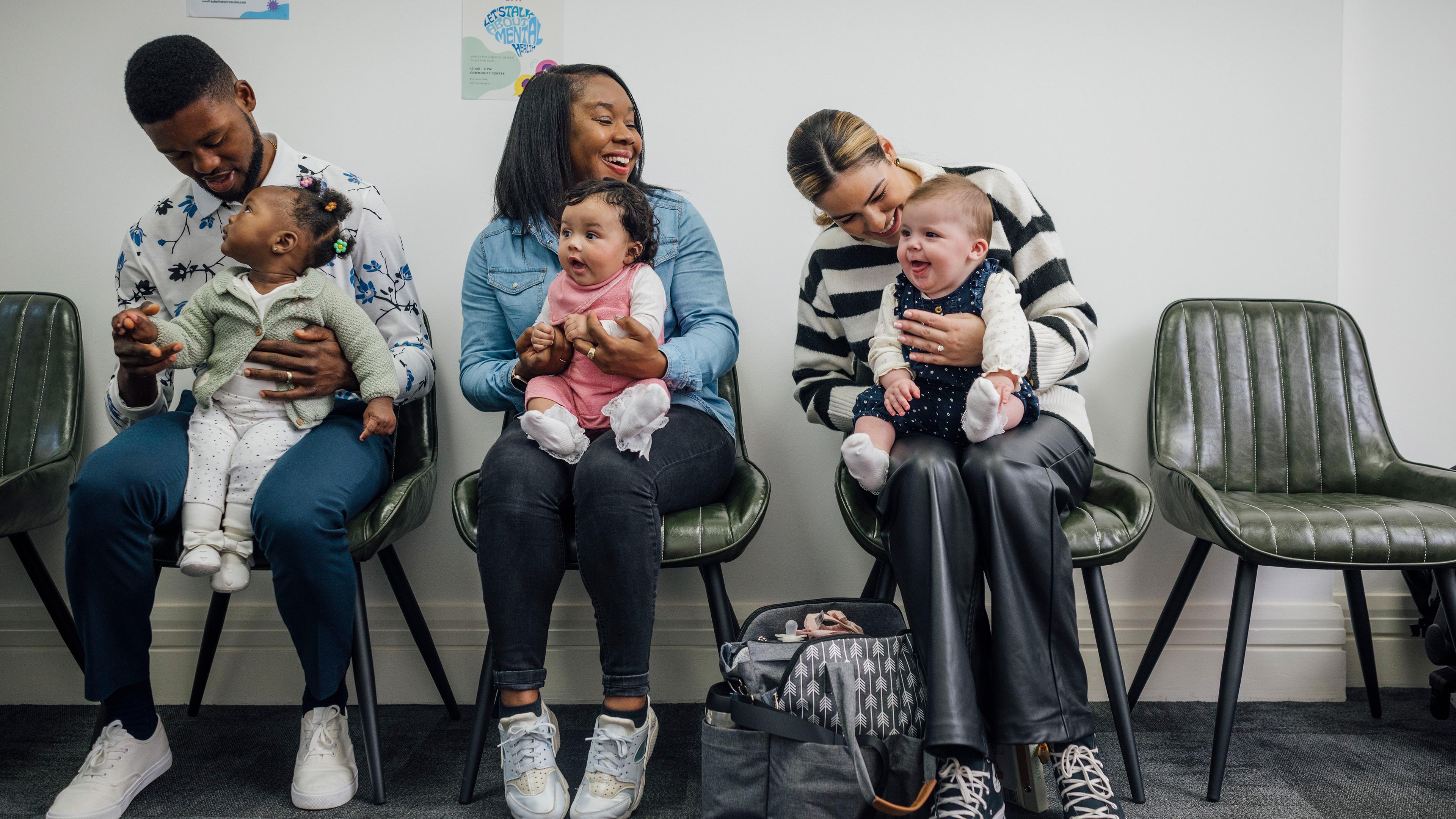 Three adults with babies seated in a waiting room, smiling and interacting, with colourful posters on the wall.