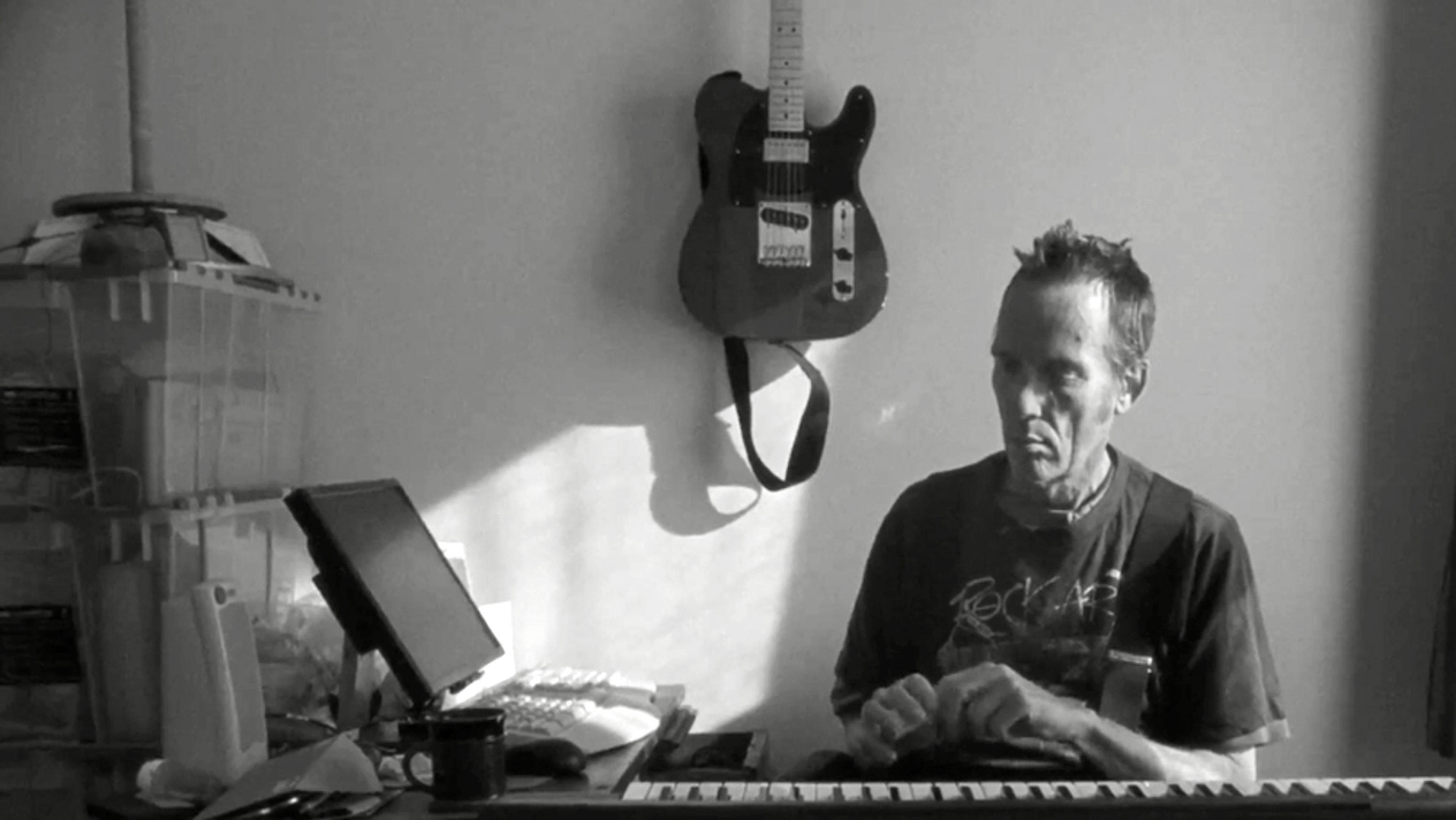 Black and white photo of a man sitting at a keyboard. A guitar hangs on the wall behind him next to a plastic storage unit.