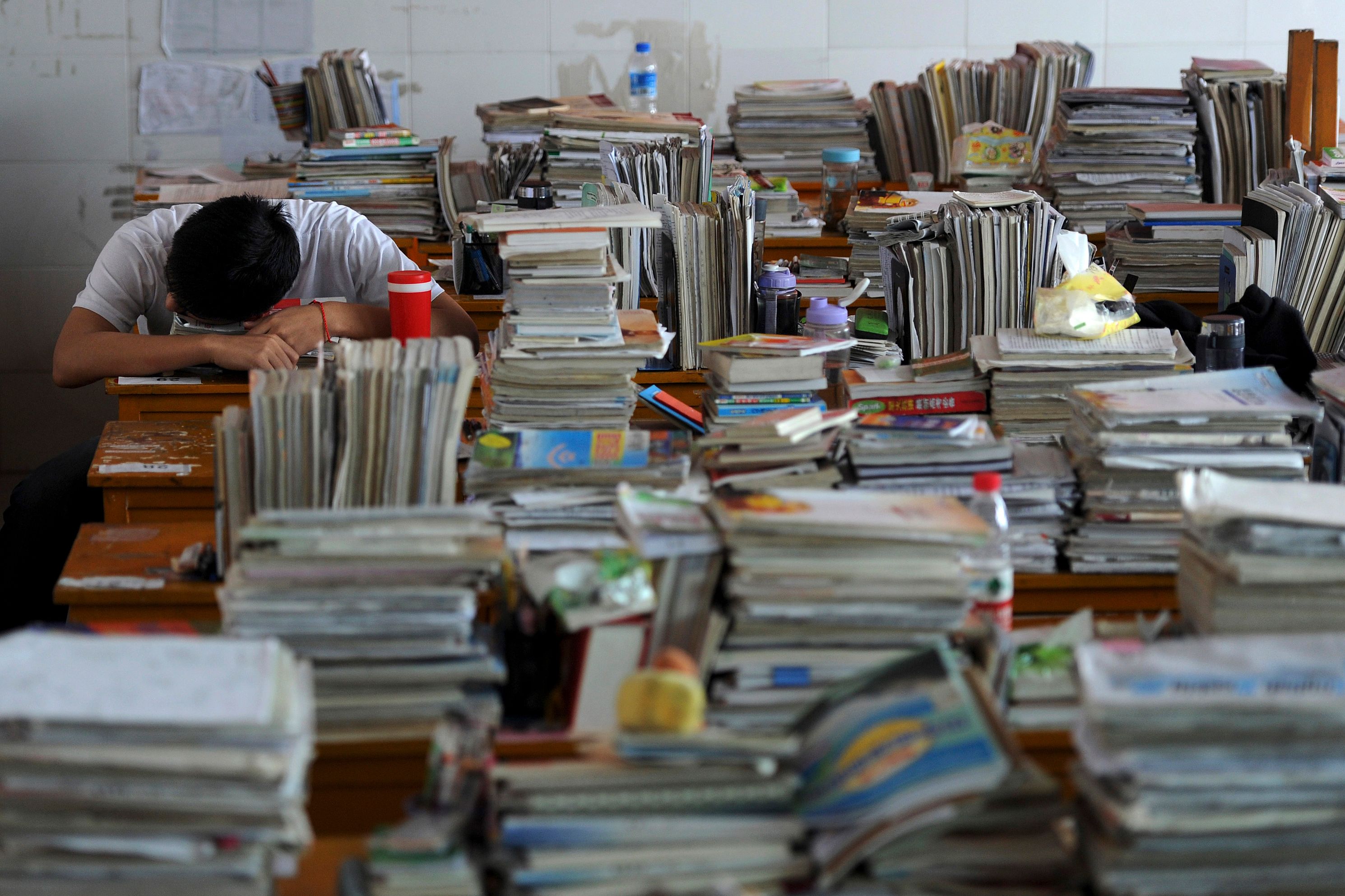 A student sleeping on a desk covered with piles of books and papers, surrounded by cluttered desks in a classroom.