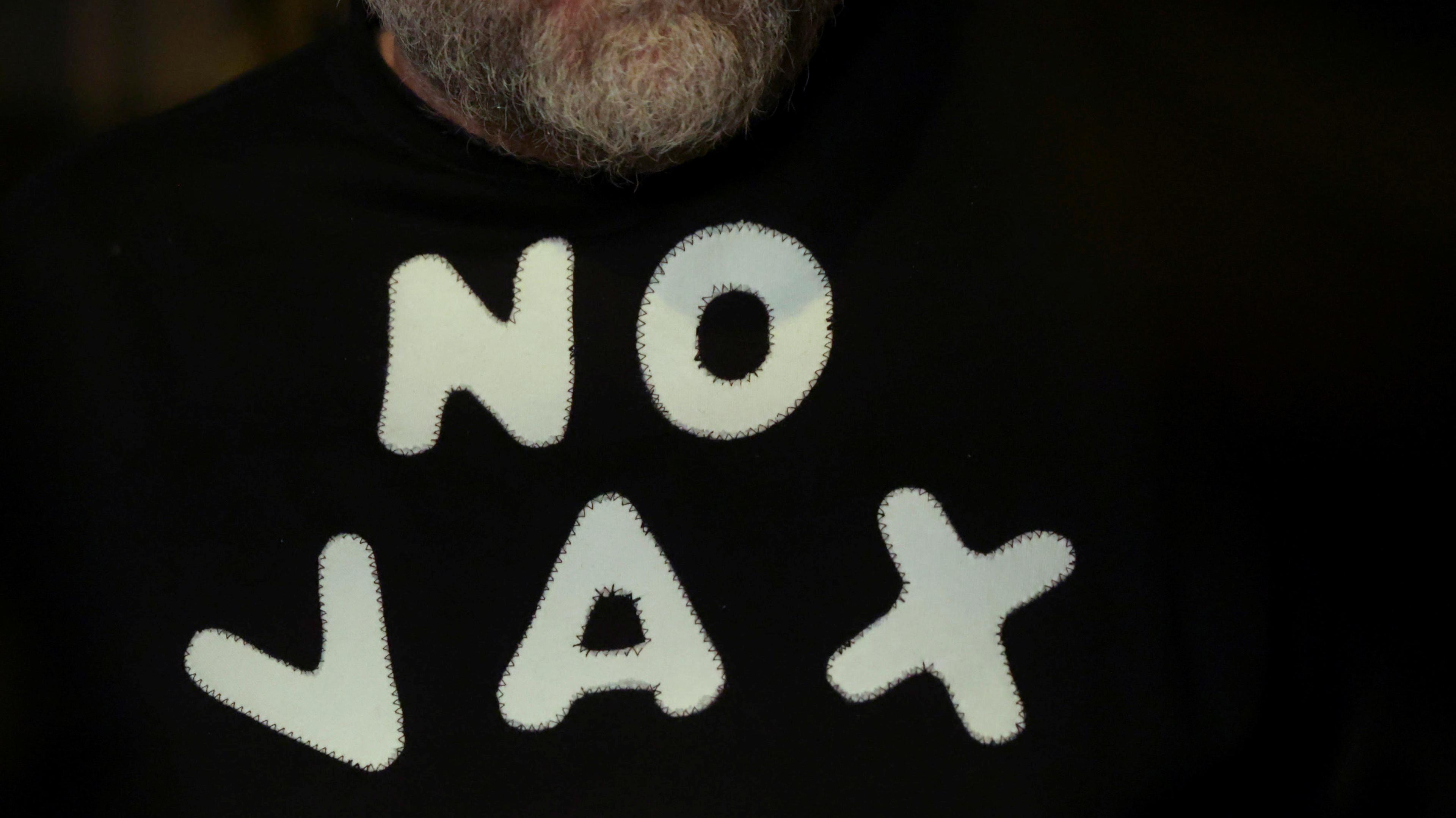 Photo of a man with a beard wearing a black shirt that has “NO VAX” in large white letters appliquéd on it.
