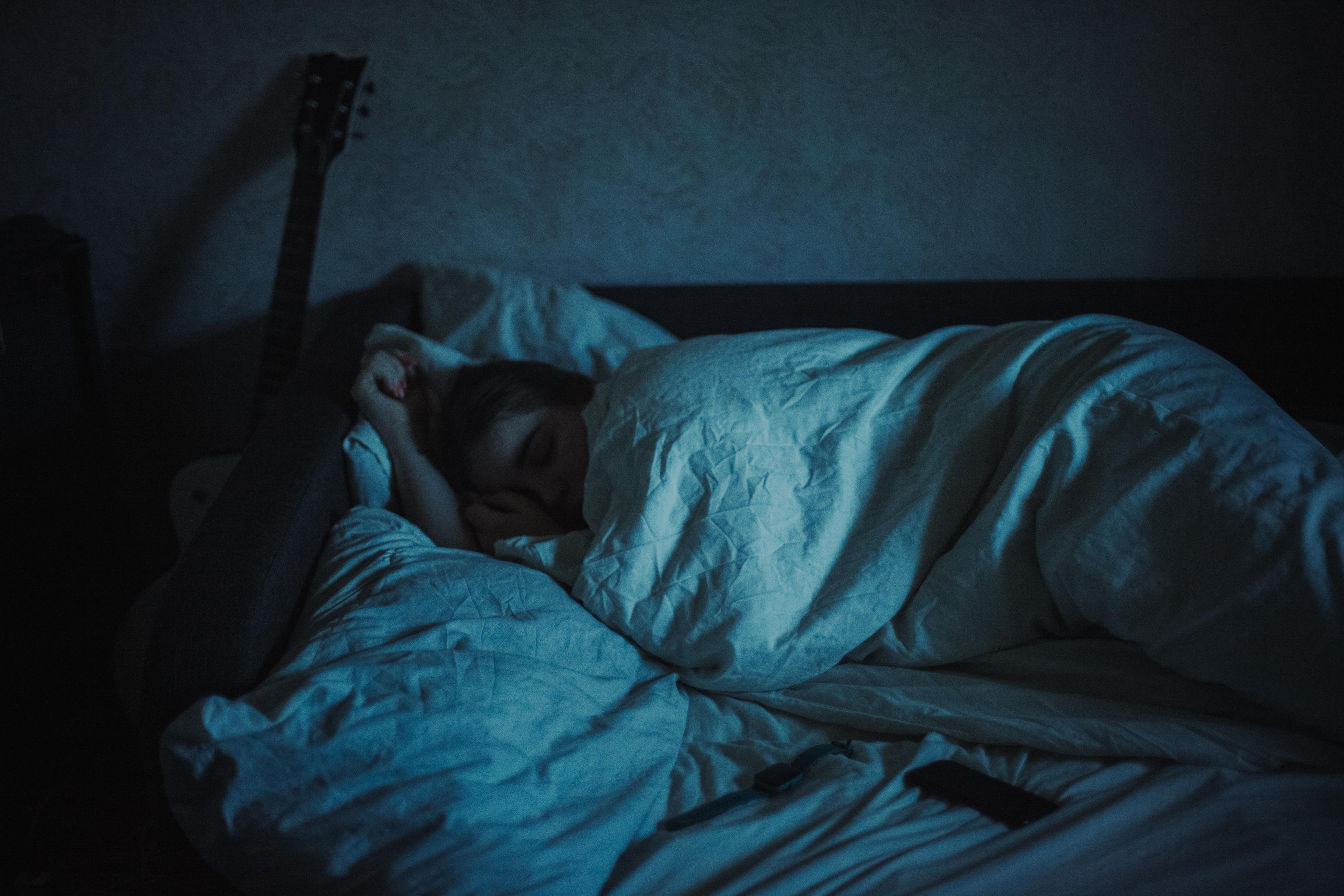 A person sleeping under a duvet on a bed with a guitar nearby, lit by dim blue light.