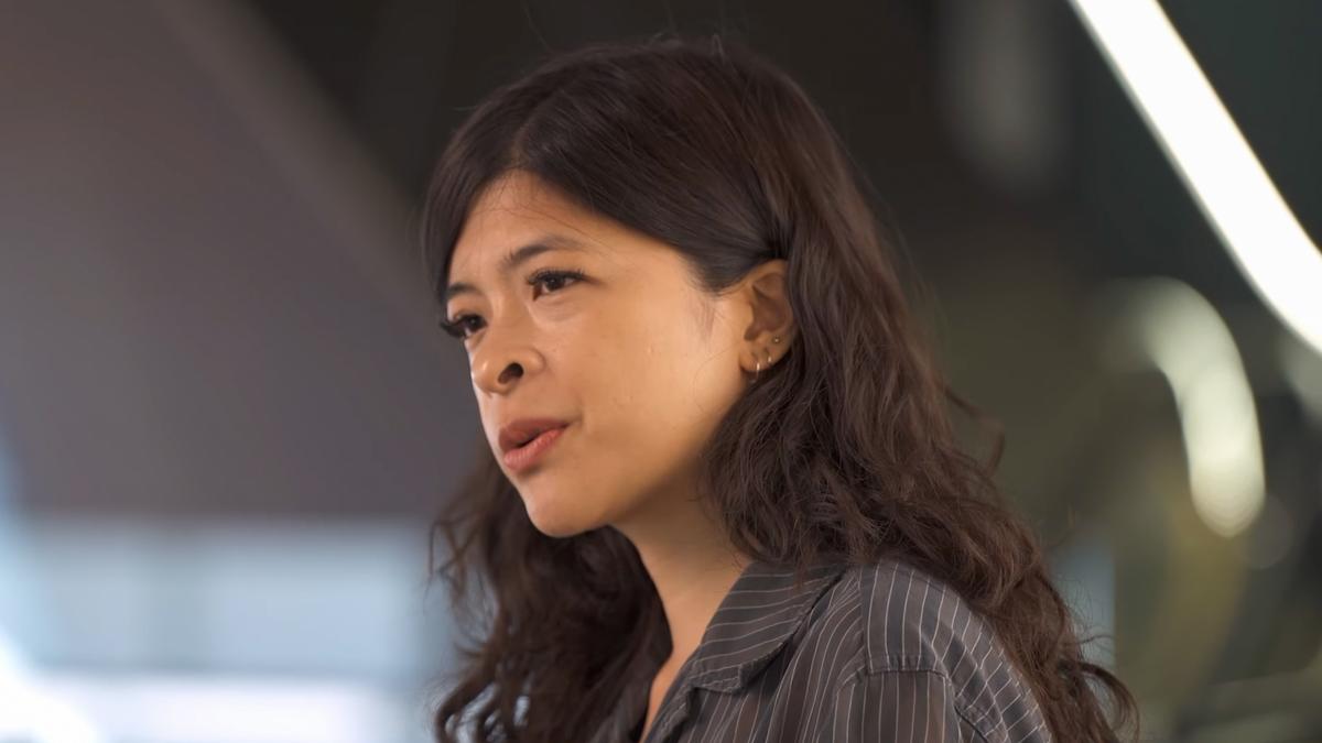 Photo of a woman with long dark hair wearing a striped shirt speaking, background blurred with soft light.
