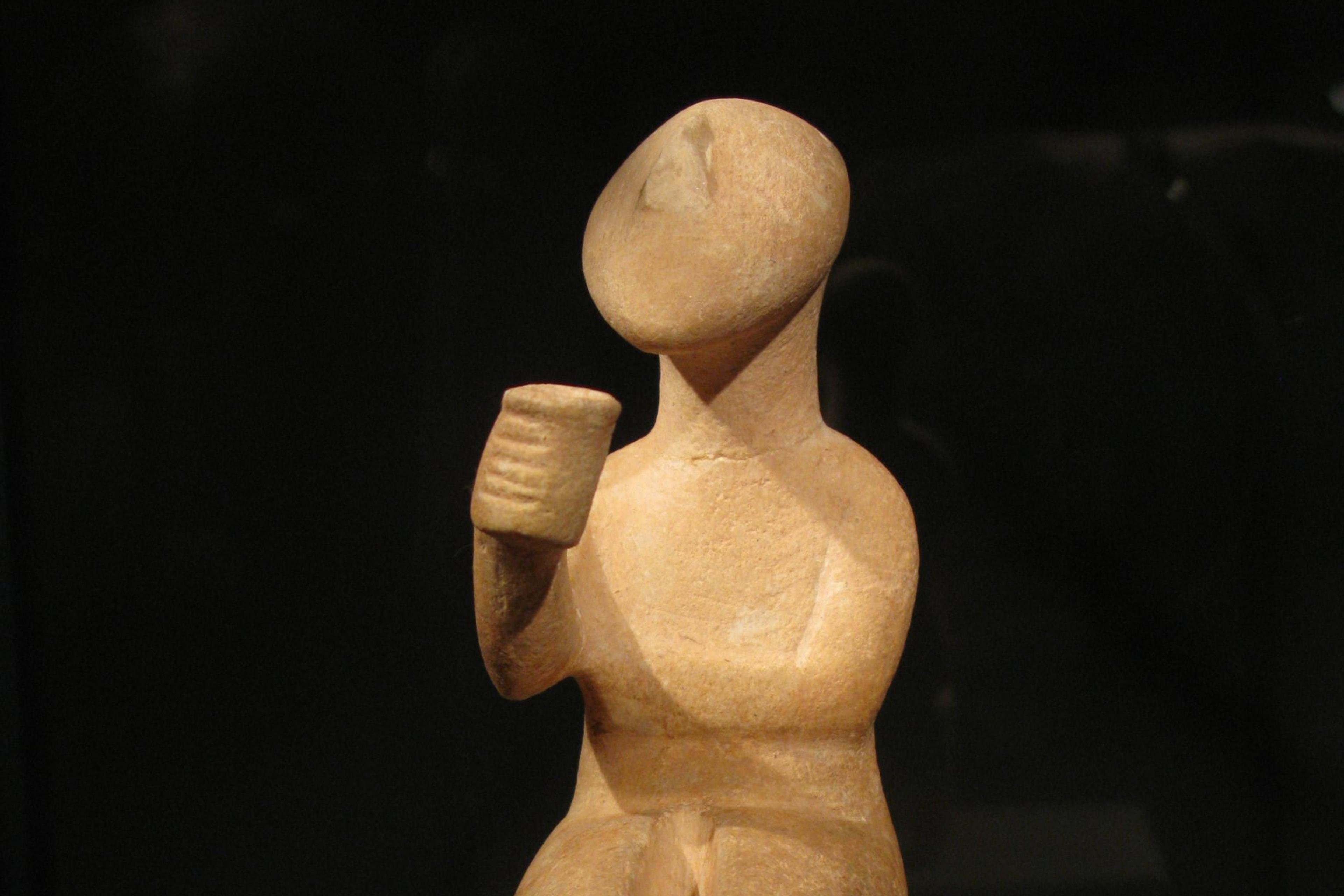 Ancient terracotta sculpture of a seated figure holding a cup, displayed against a dark background.