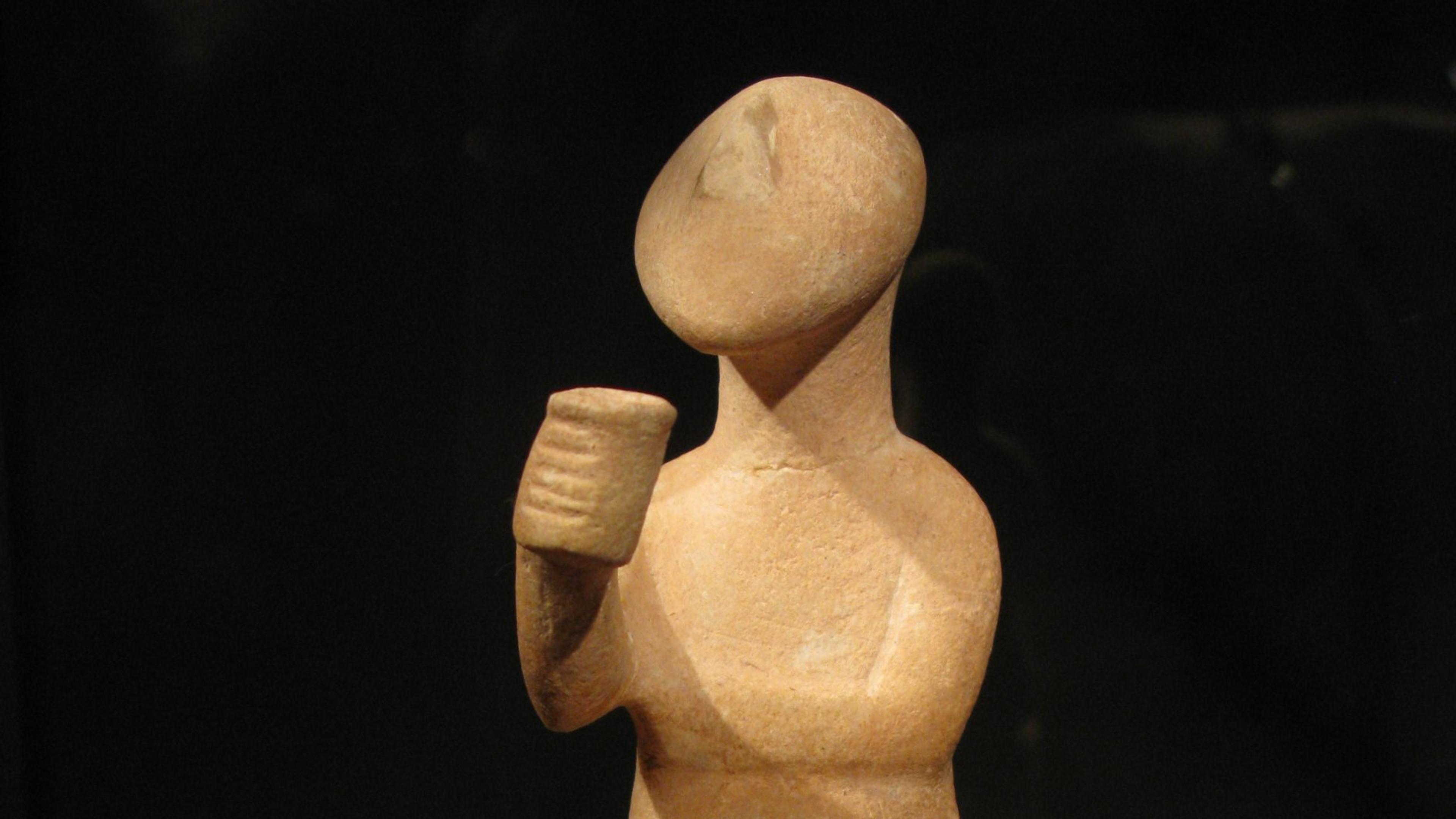 Ancient terracotta sculpture of a seated figure holding a cup, displayed against a dark background.