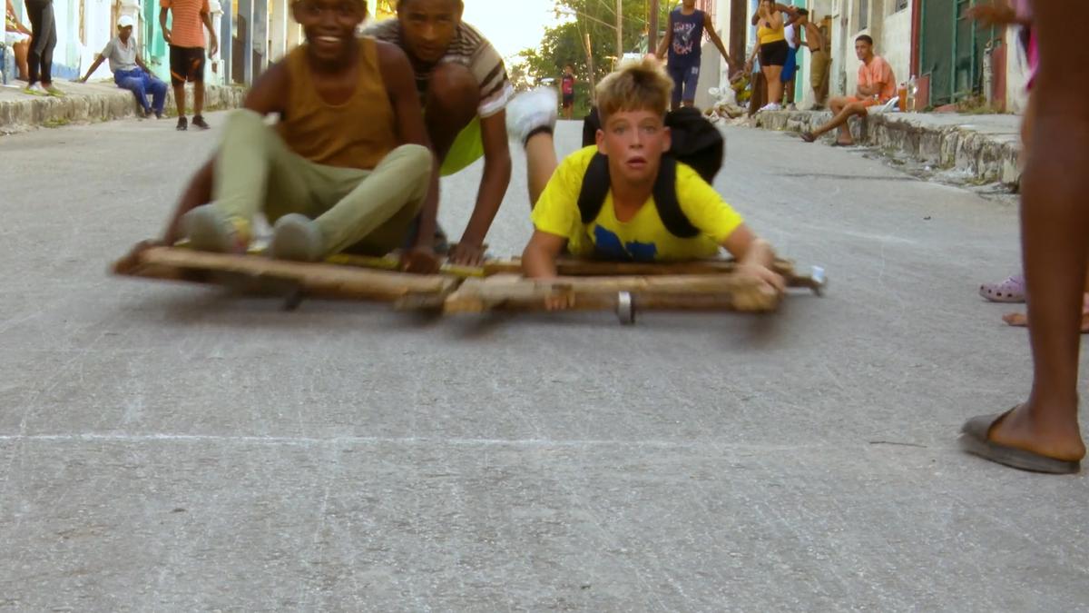 Photo of three boys riding wooden carts down a street in an urban setting, with onlookers in the background.