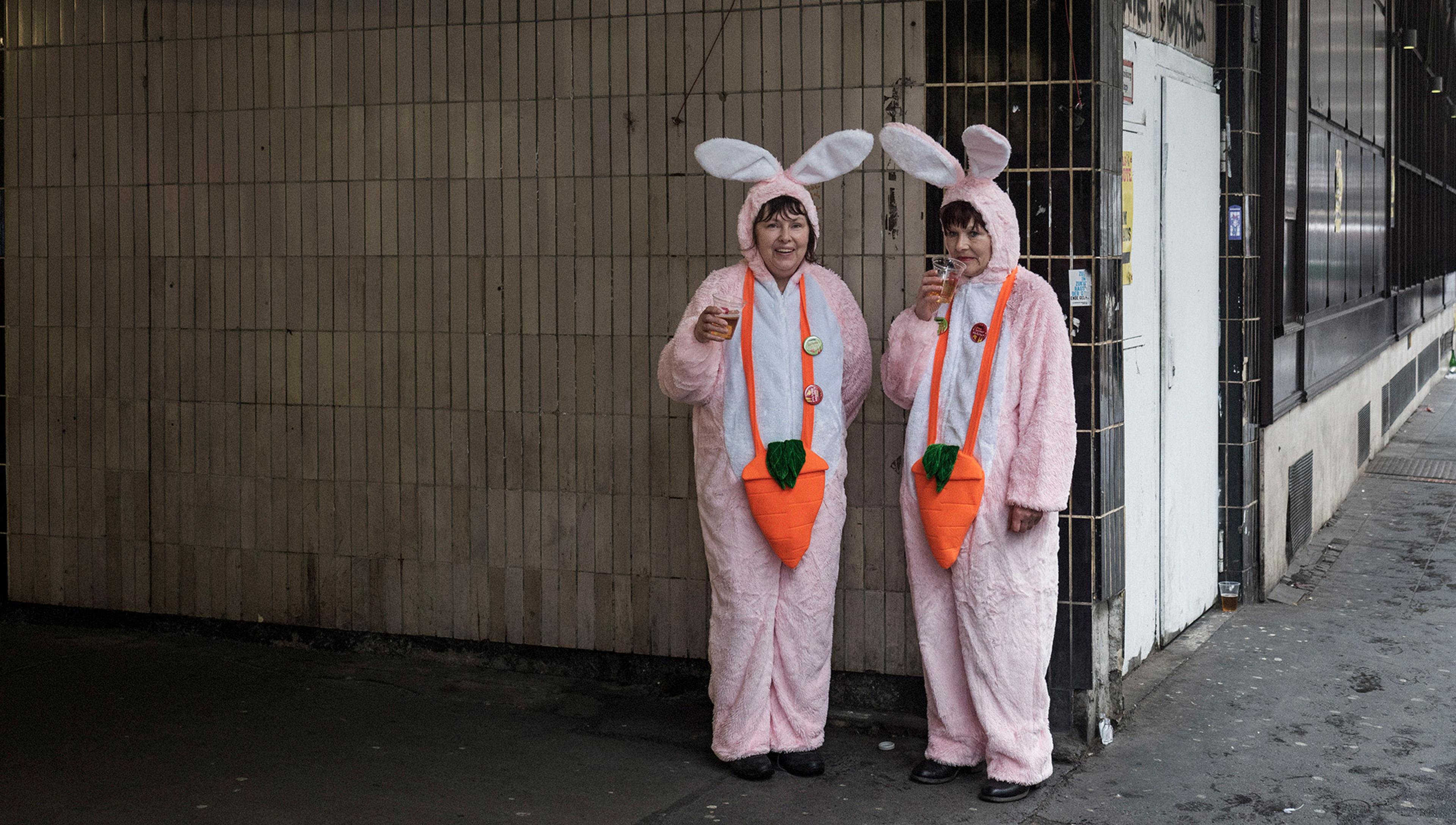 Two women dressed in pink and white rabbit outifts drink a beer outside a subway station entrance. One of them looks glum
