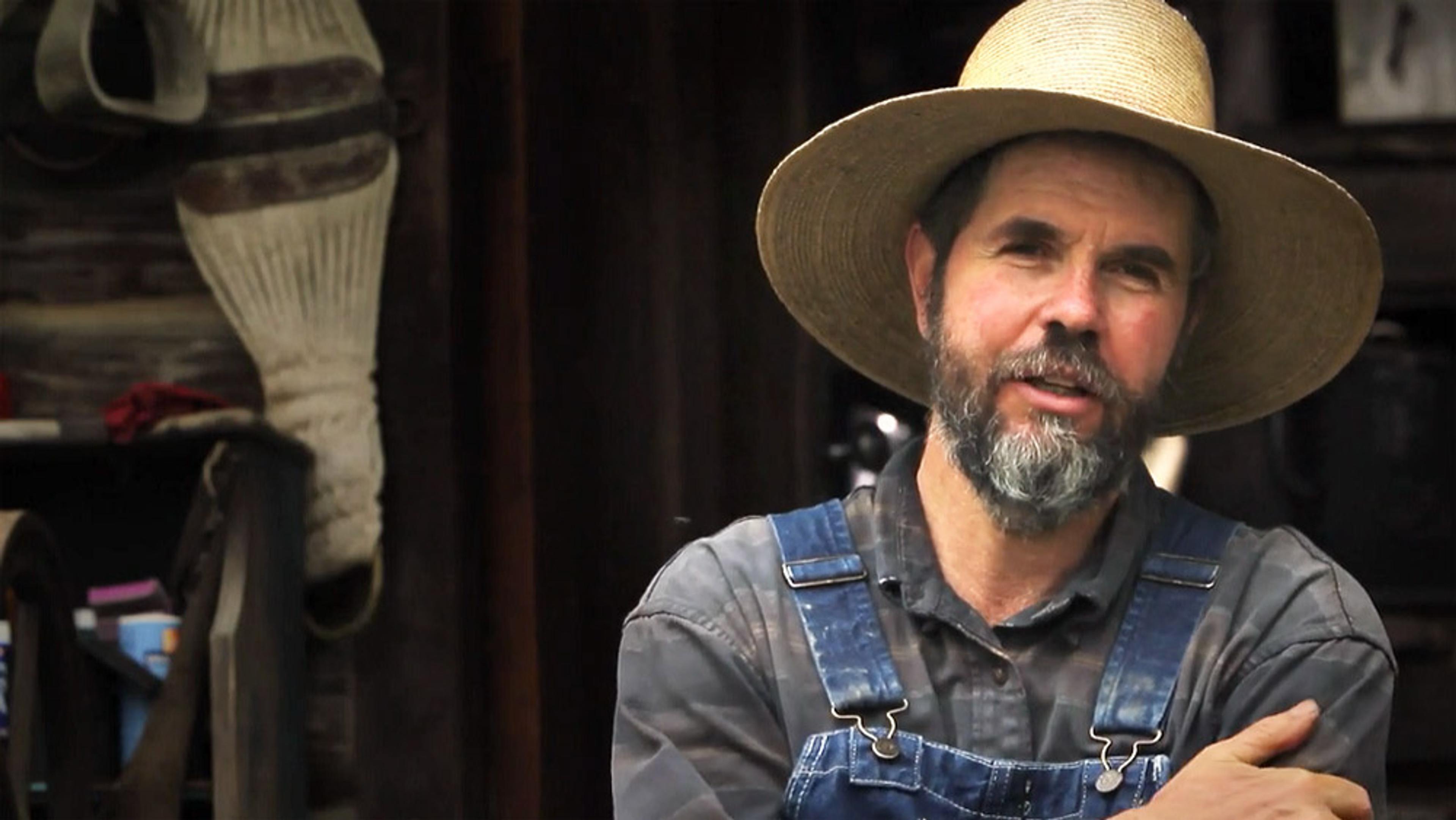 A bearded man wearing a straw hat and denim overalls, standing with his arms crossed in front of a rustic background.