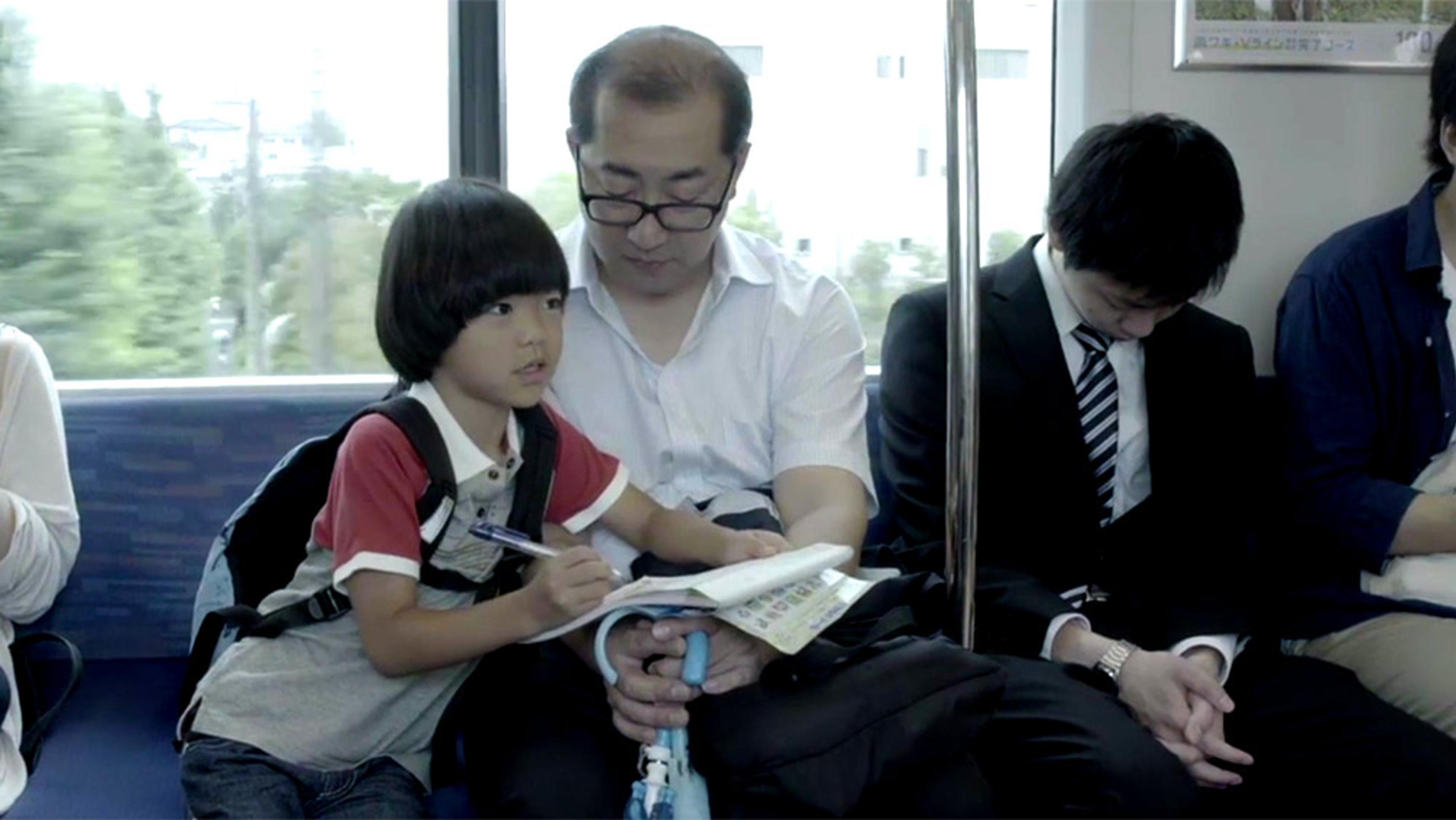 A child writing in a notebook on a train, next to a man with glasses reading and a man in a suit sleeping.