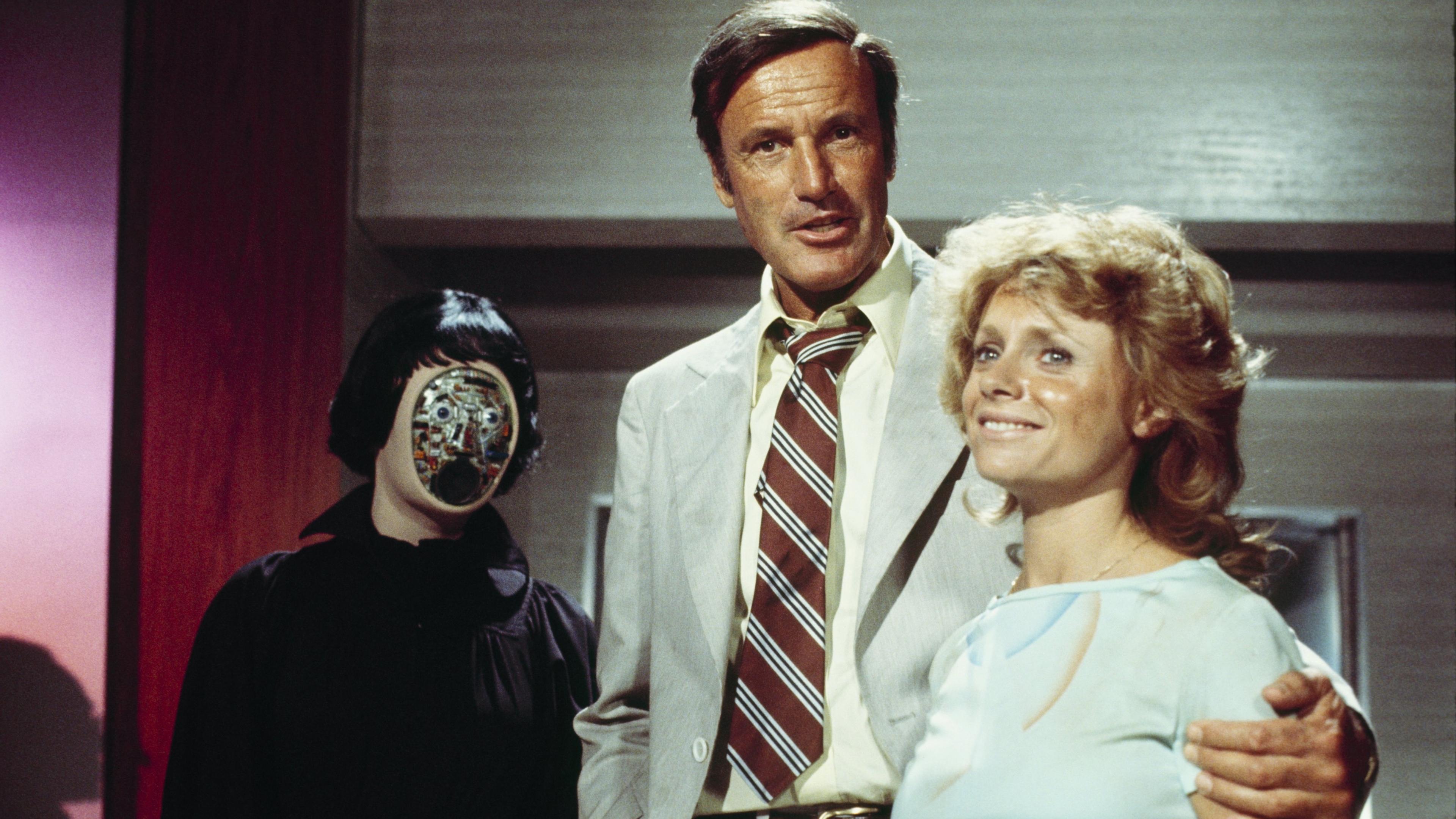 Photo of a man in a suit with two people, one with a mechanical face, indoors.