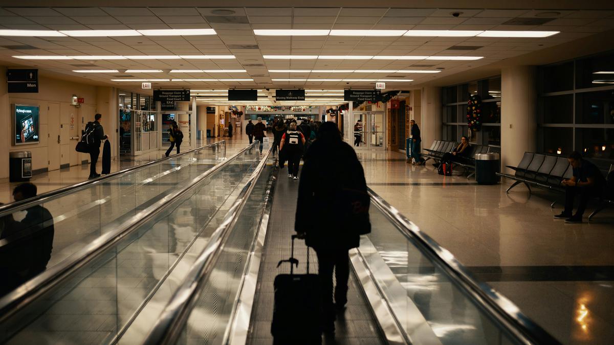 Airport terminal with people on moving walkway pulling luggage and others sitting on benches.