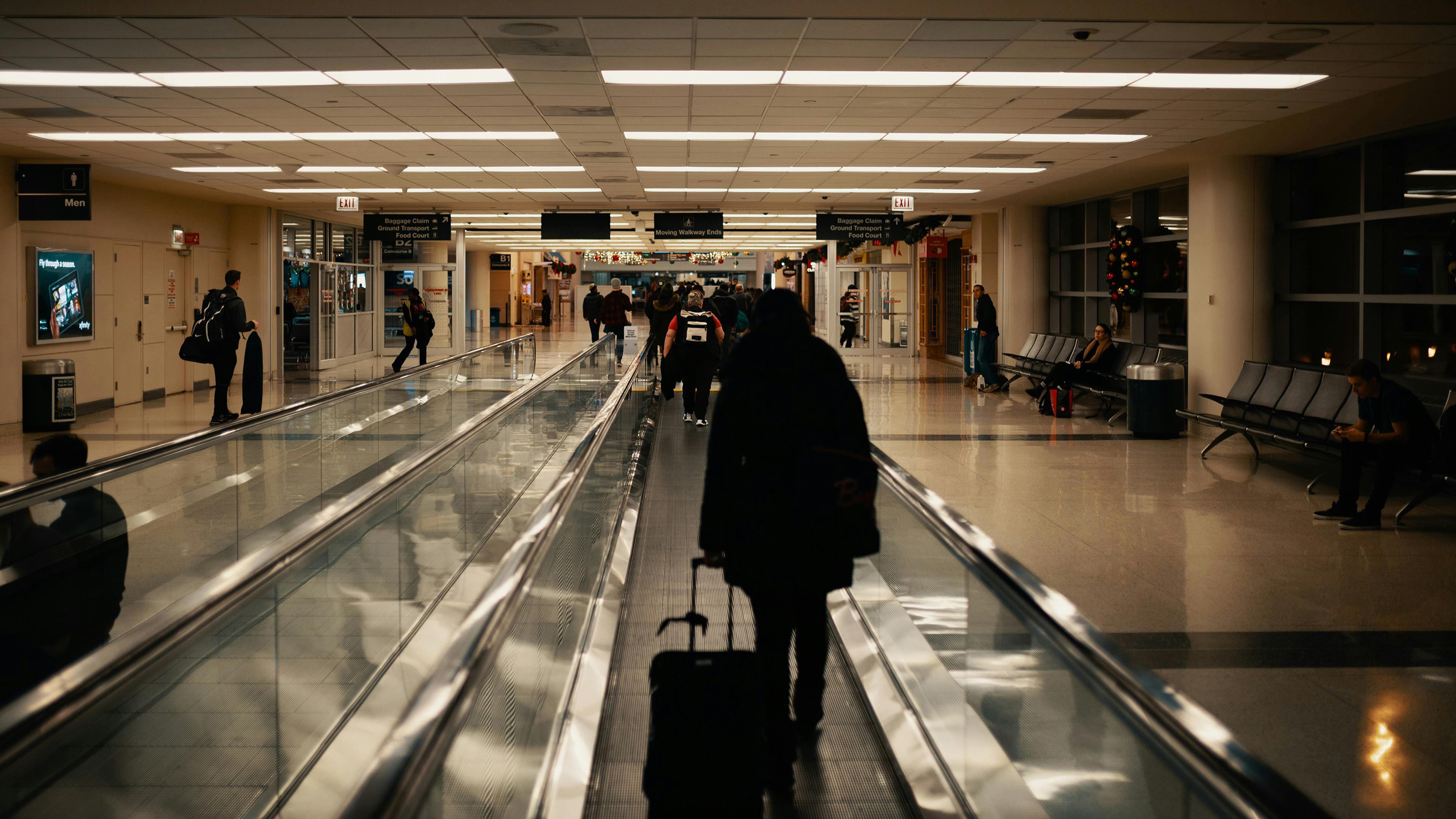 Airport terminal with people on moving walkway pulling luggage and others sitting on benches.
