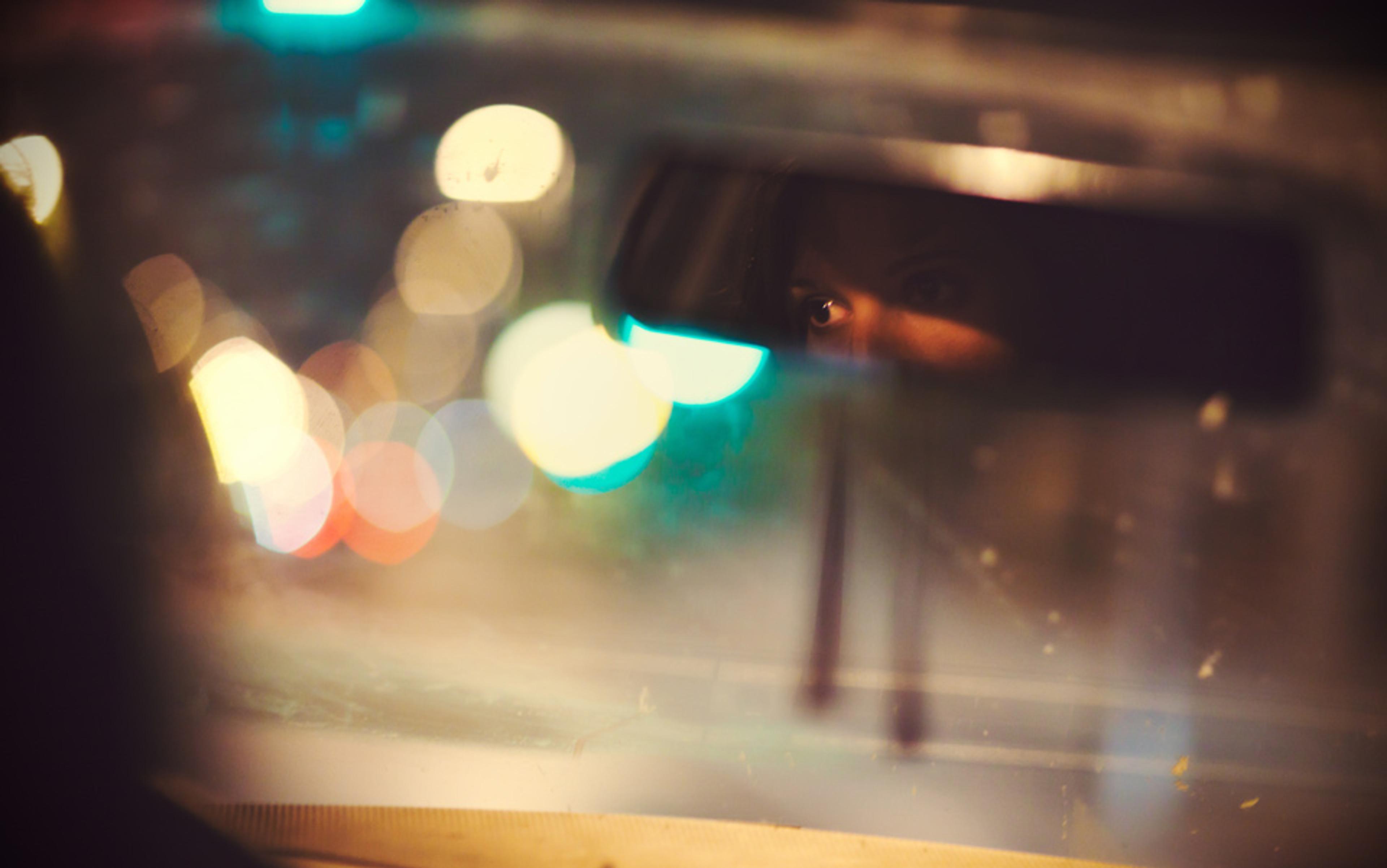 A car’s rear-view mirror showing a person’s eyes, with blurry city lights visible through the windshield at night.