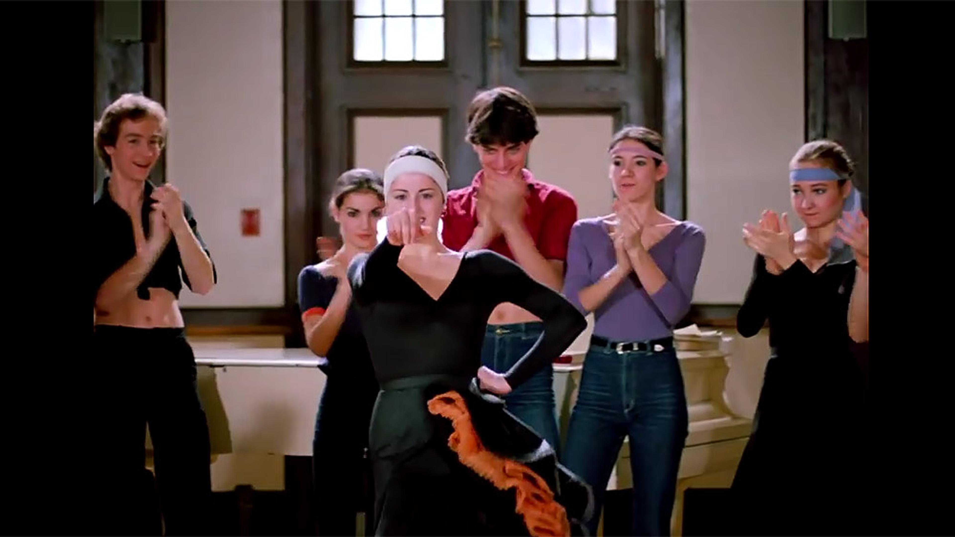 A woman in black dancewear performs with dancers clapping supportively in the background.