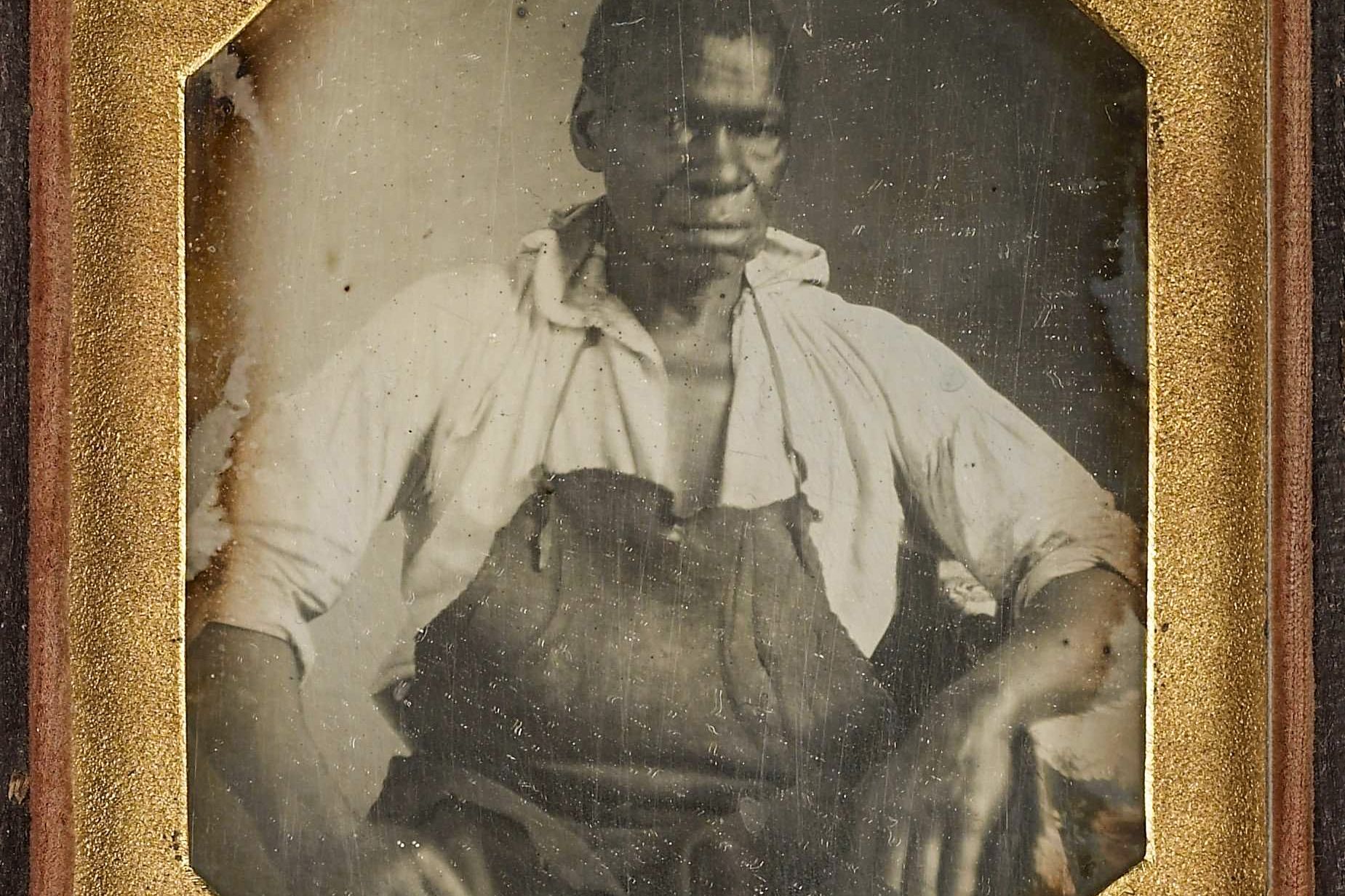 Daguerreotype of a seated man wearing an apron and a partially open shirt framed in a gold mat.
