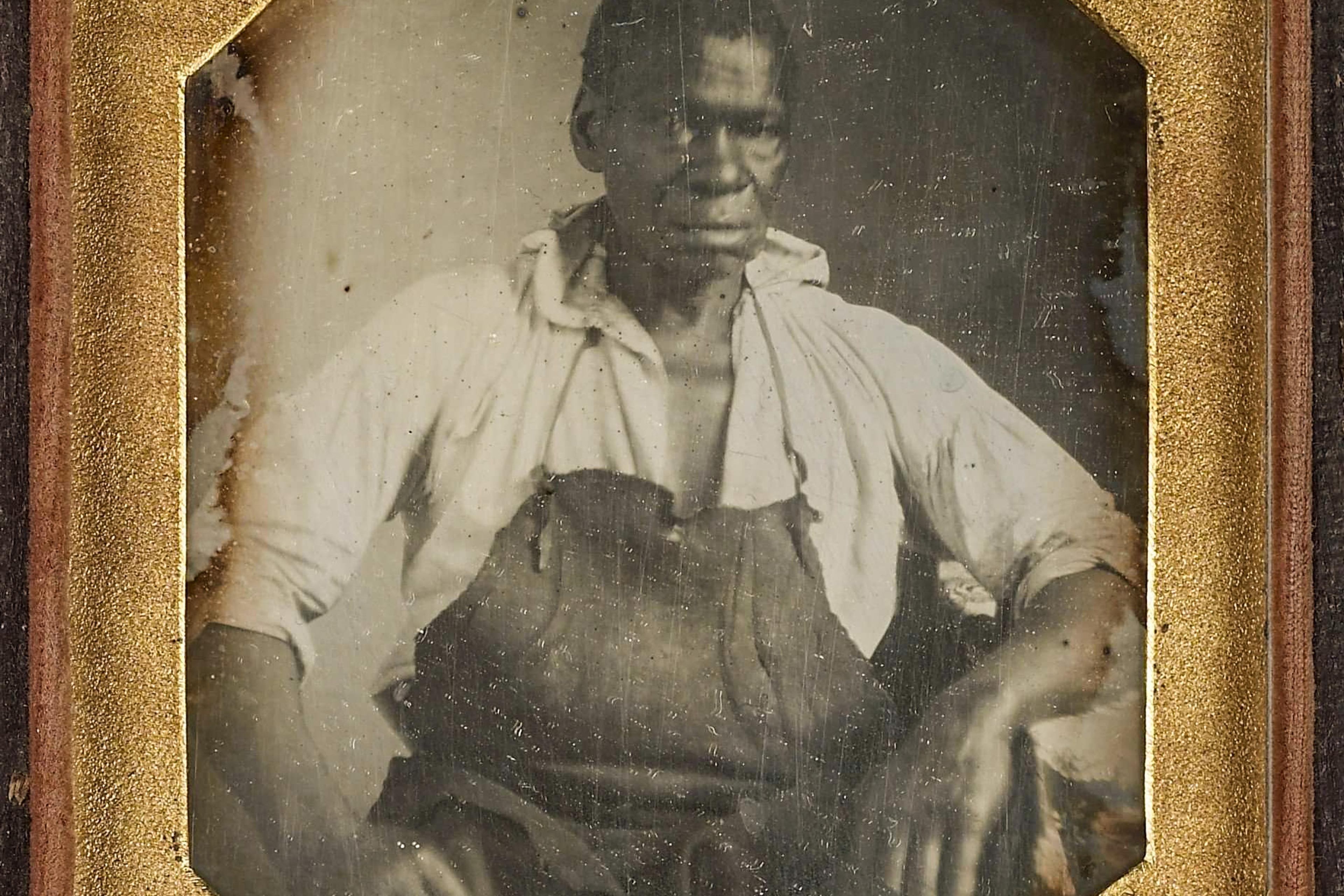 Daguerreotype of a seated man wearing an apron and a partially open shirt framed in a gold mat.