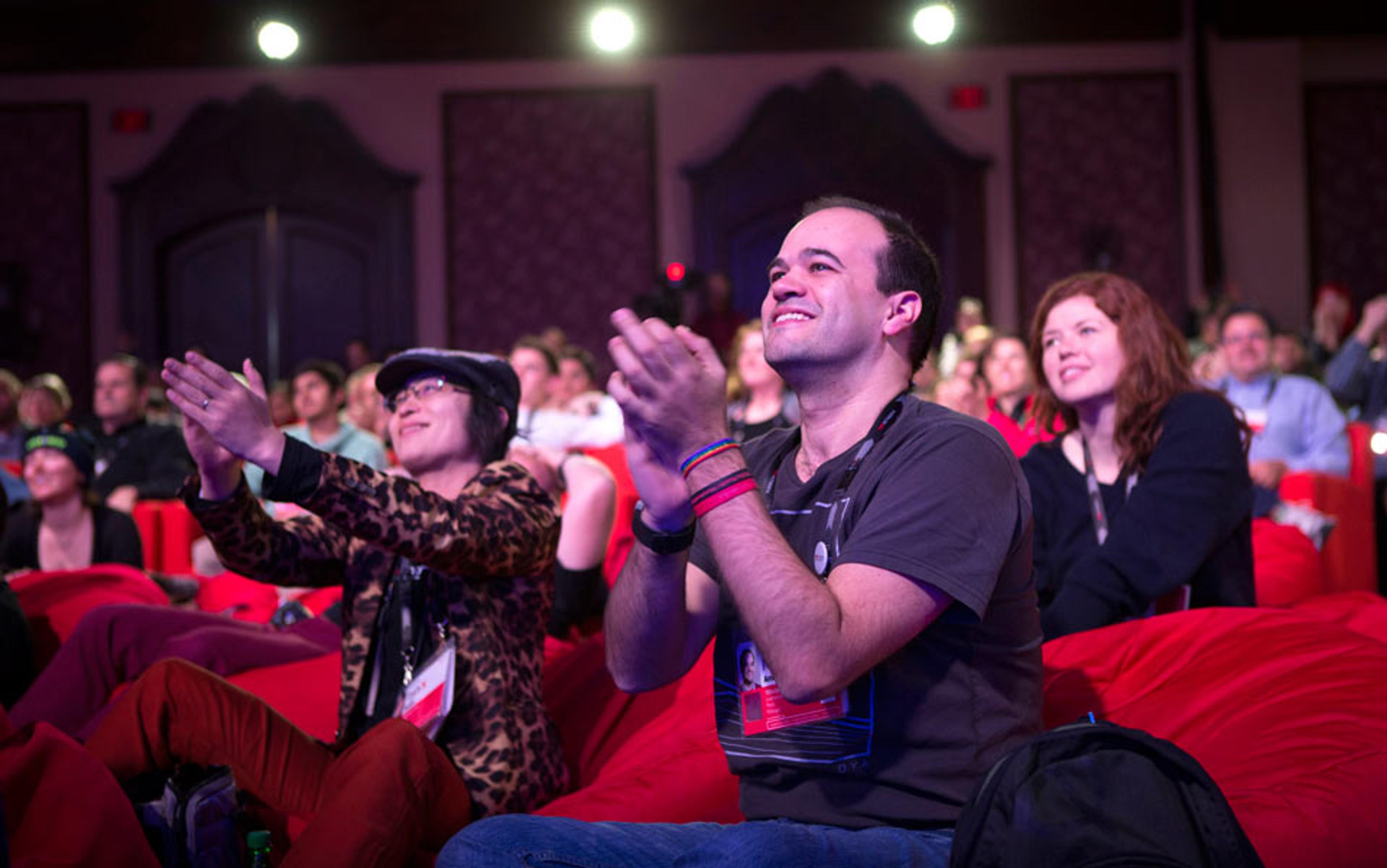 A happy audience at a conference seated on red beanbags clapping and smiling with decorative walls and lights in the background.