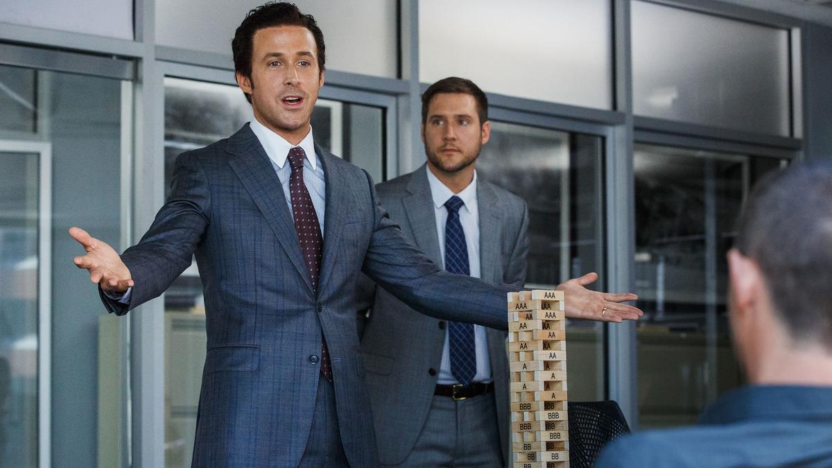 Photo of a businessman in a suit explaining with a Jenga tower, another man stands behind him, both in an office setting.
