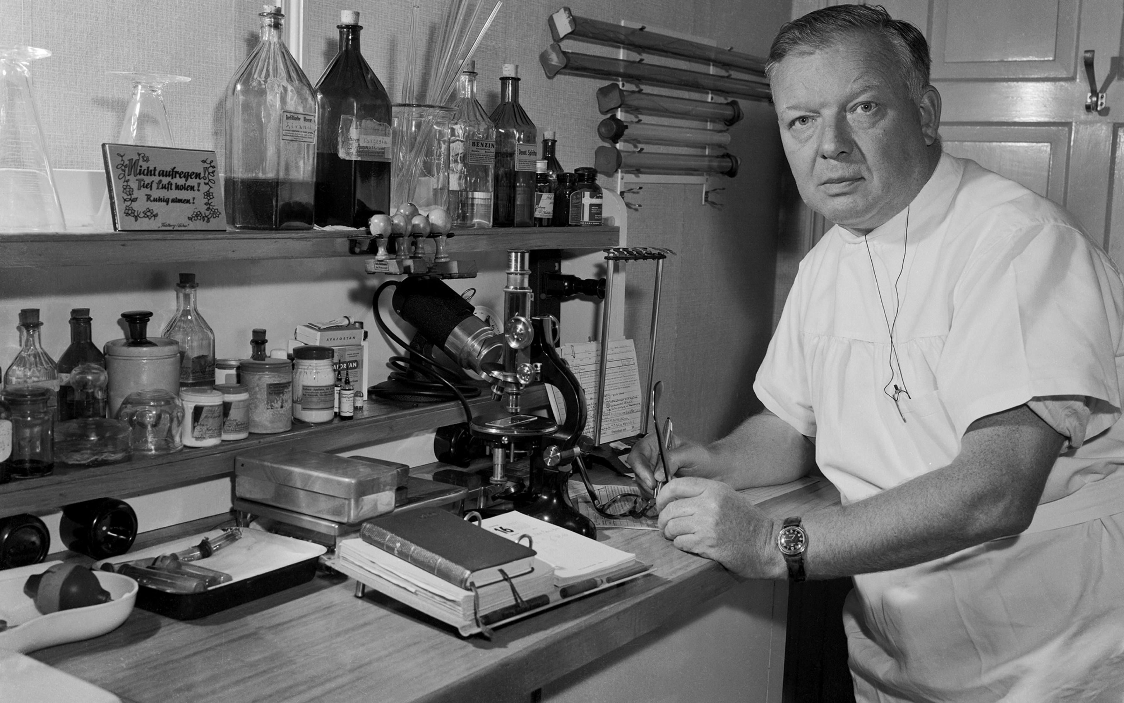 A black and white photo of a man in doctor’s clothing posed next to a lab bench with a microscope looking directly at the camera