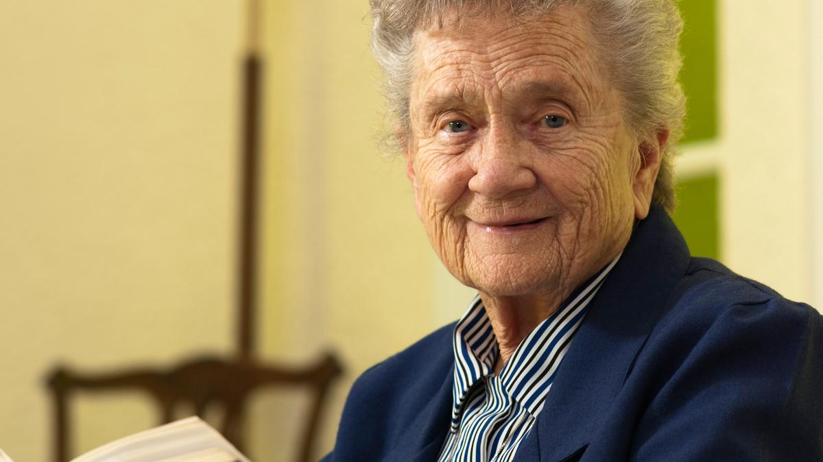 An elderly woman in a blue jacket holding a book, smiling warmly at the camera indoors.