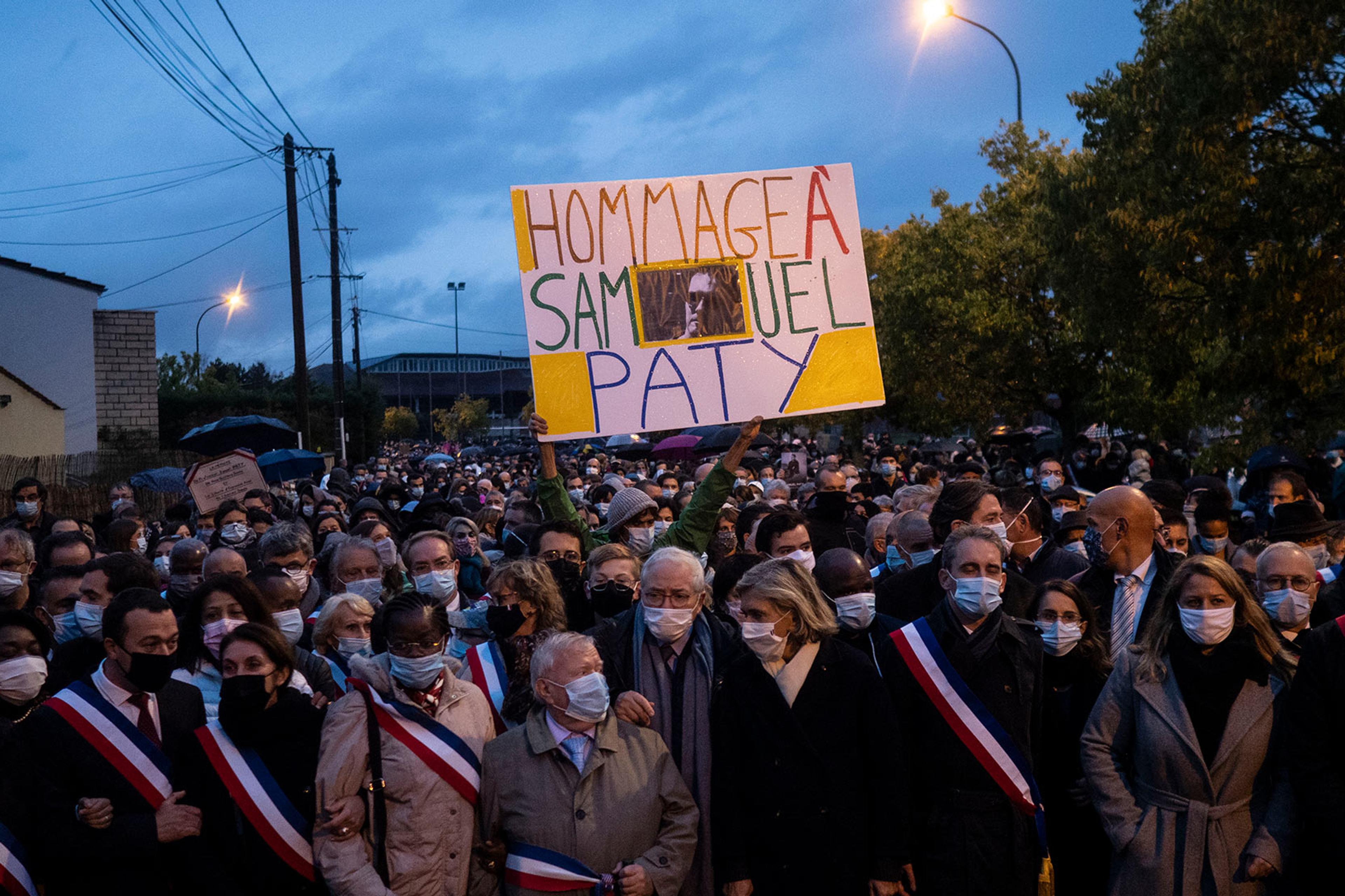 A large crowd wearing masks and sashes, holding a sign reading “Hommage à Samuel Paty” against an evening sky.