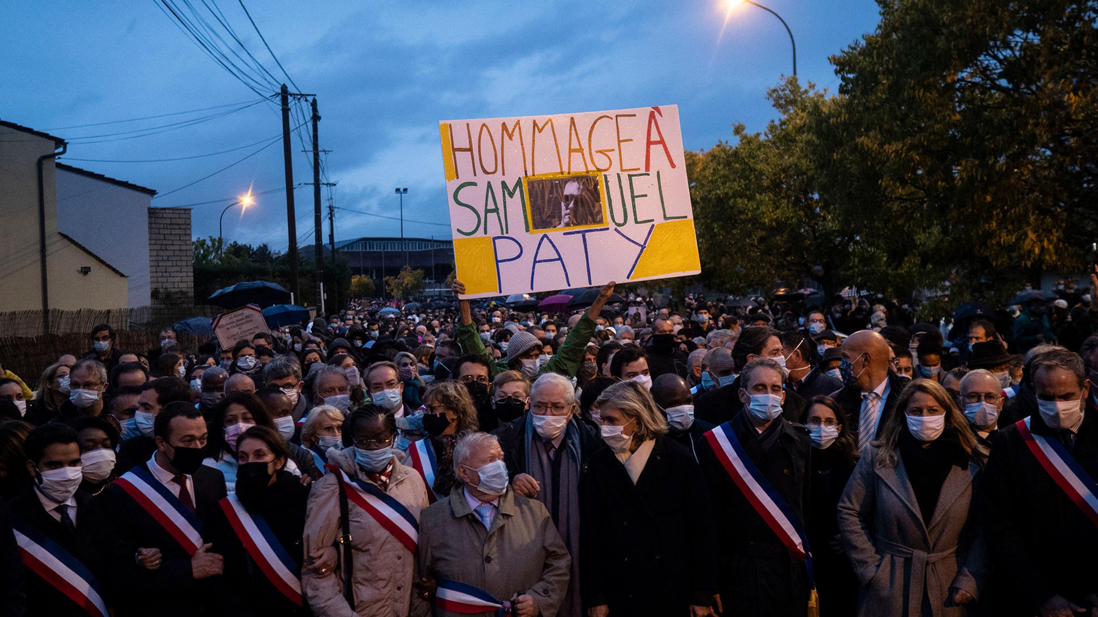 A large crowd wearing masks and sashes, holding a sign reading “Hommage à Samuel Paty” against an evening sky.