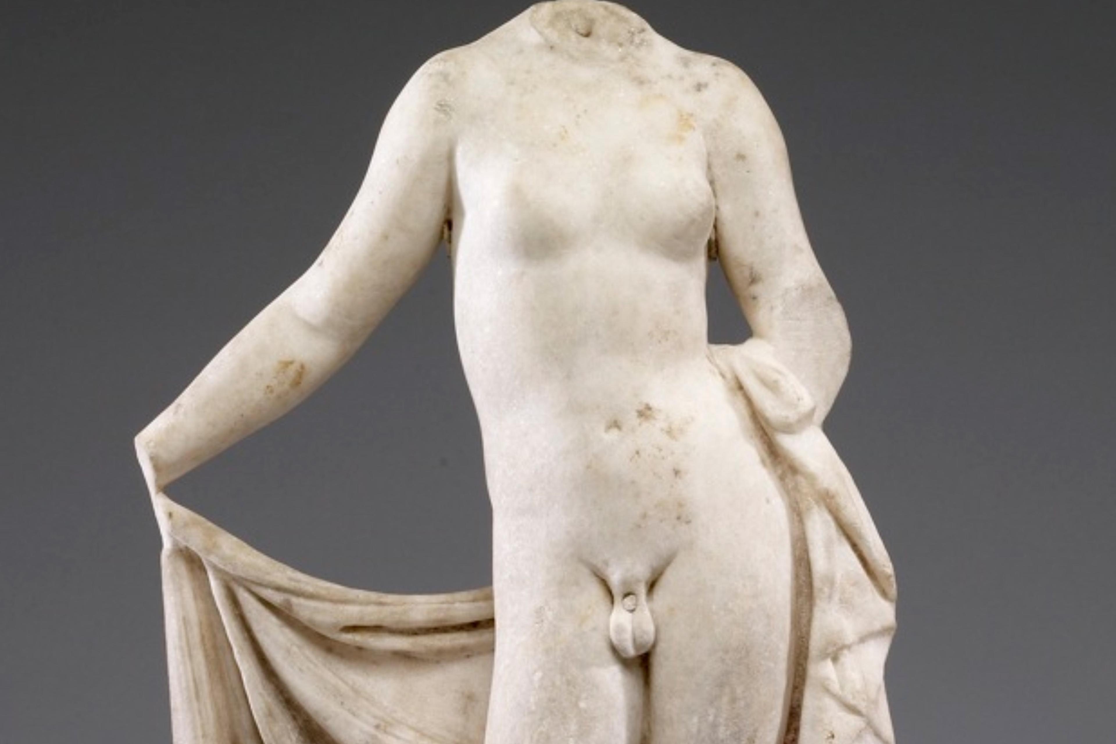 Ancient marble statue of a headless Hermaphroditus, draped lower garment, against a plain background.