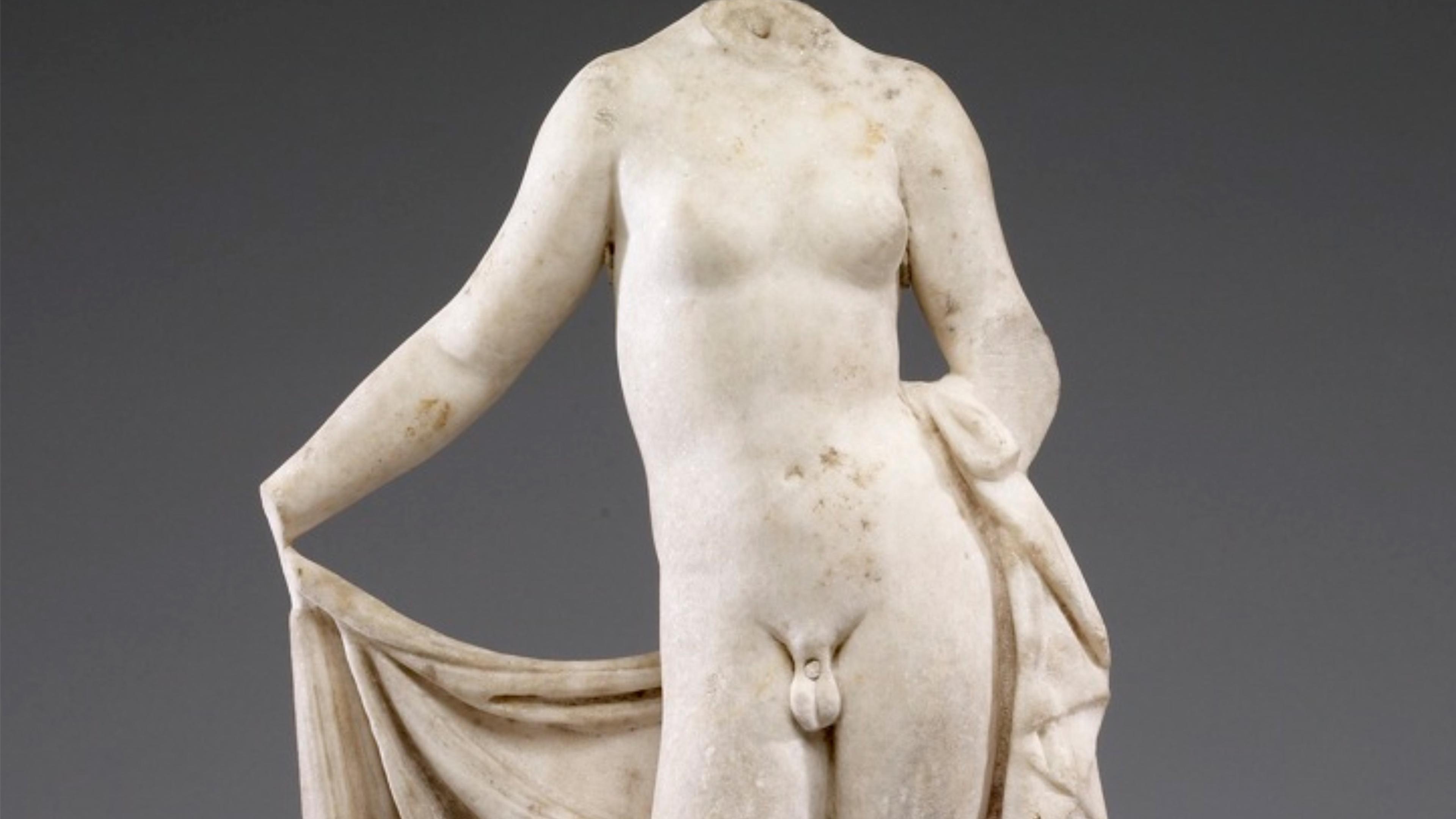 Ancient marble statue of a headless Hermaphroditus, draped lower garment, against a plain background.