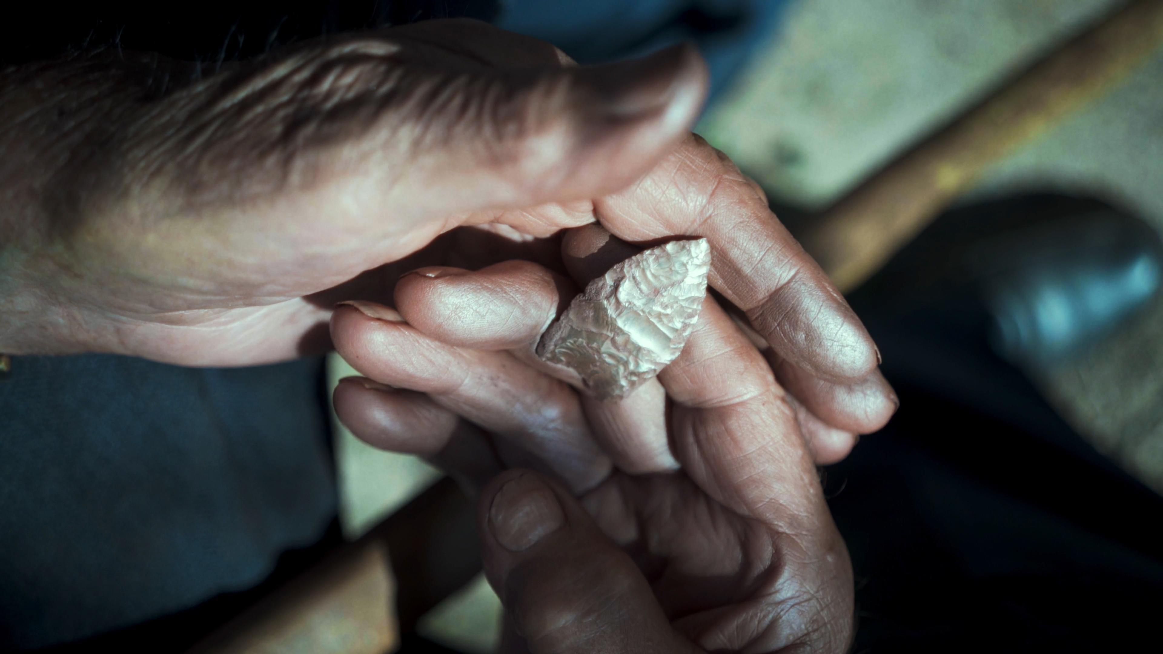 Weathered hands holding a small, carved stone tool with intricate details, set against a blurred background.