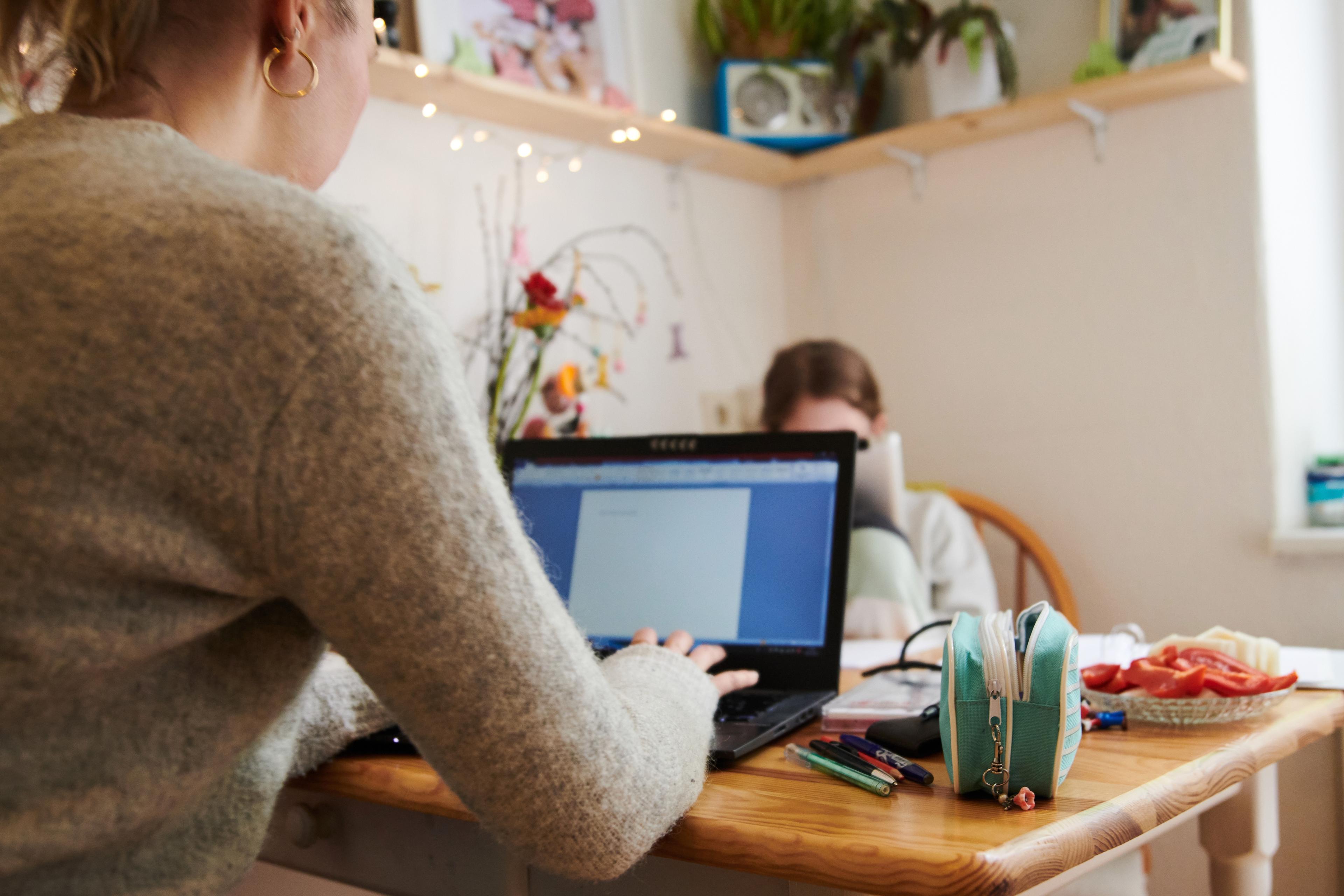 Photo of a person using a laptop on a wooden table with stationery books and a plate of snacks also visible.