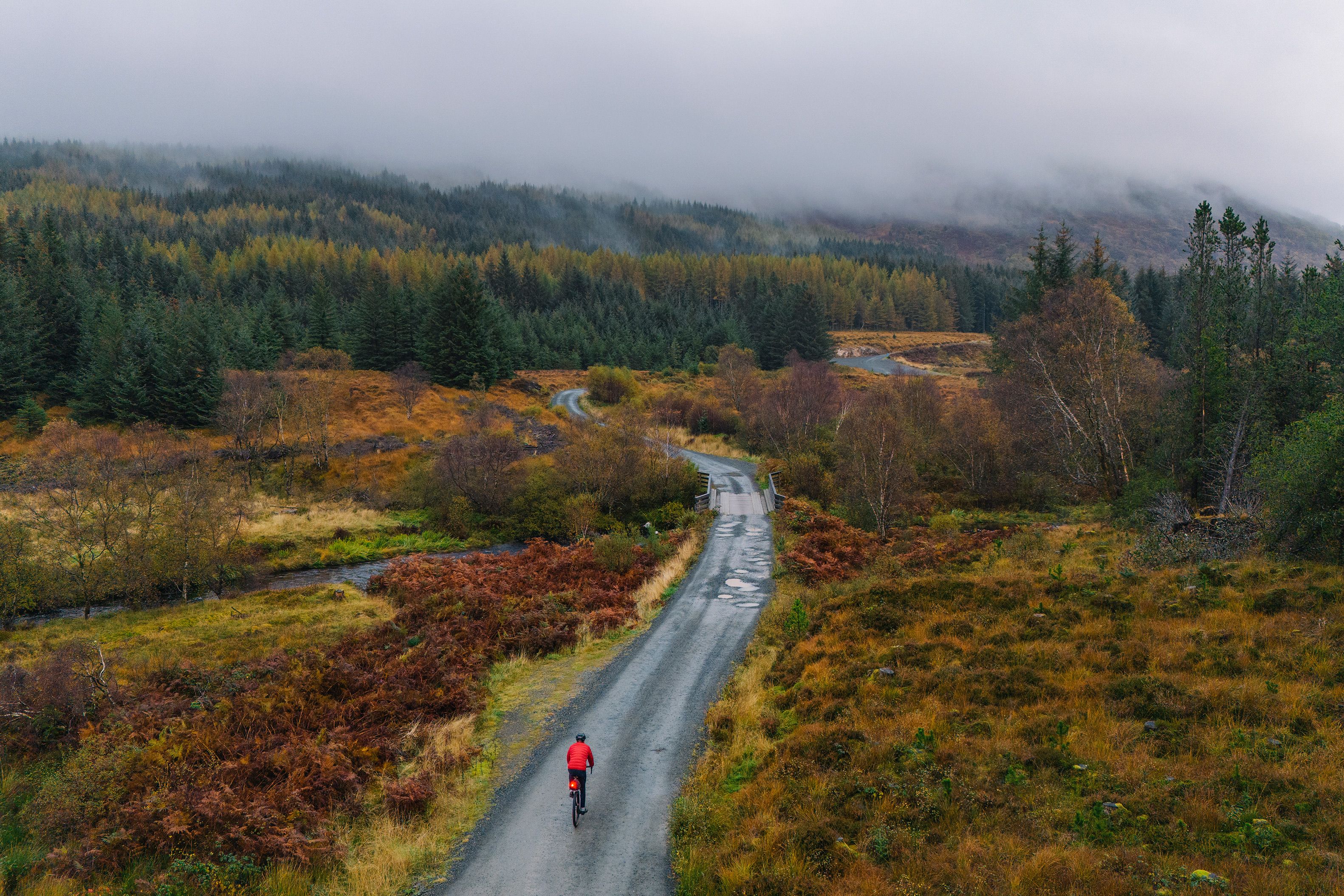 A cyclist in a red jacket on a winding rural road surrounded by autumnal trees with misty hills in the background.