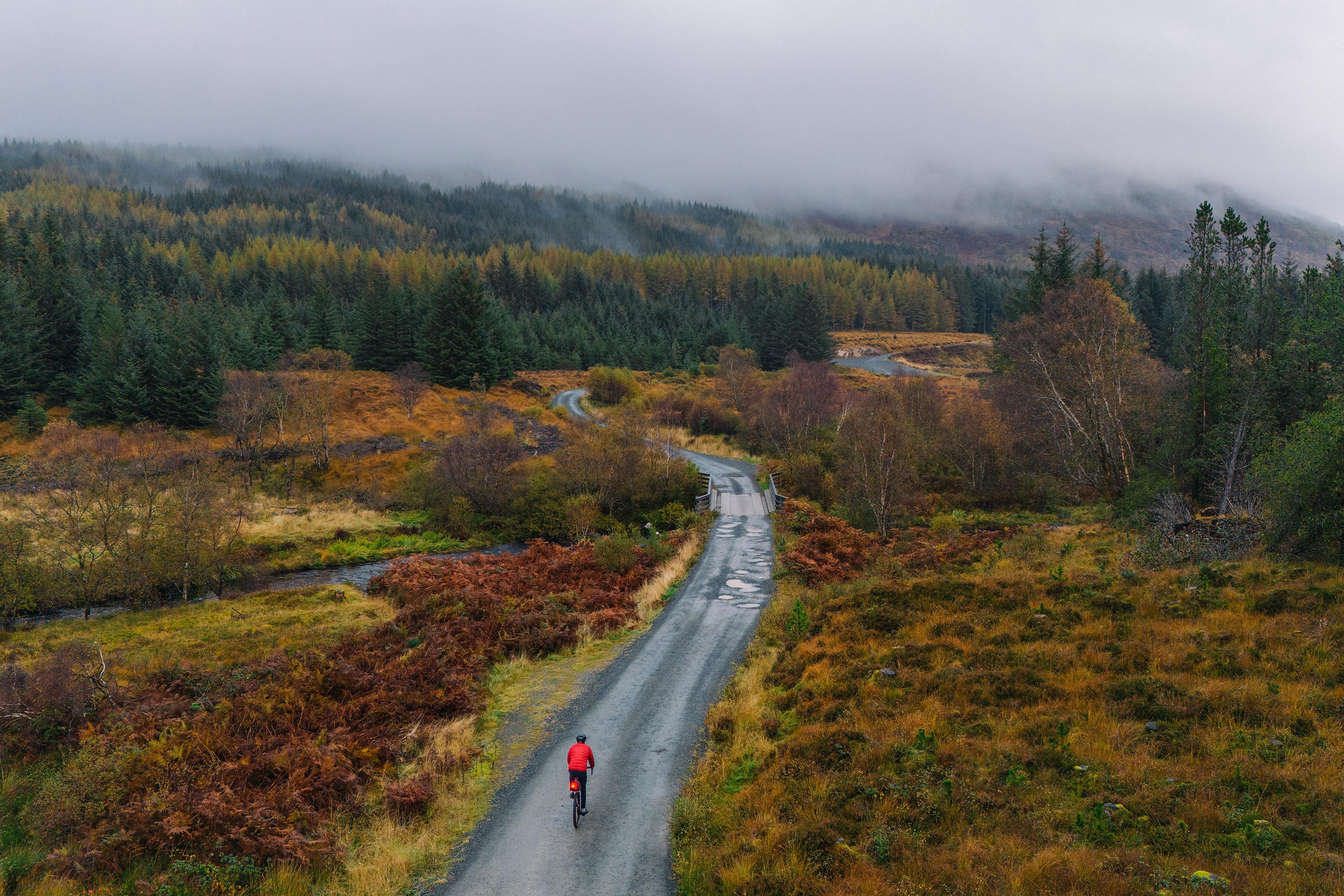 A cyclist in a red jacket on a winding rural road surrounded by autumnal trees with misty hills in the background.