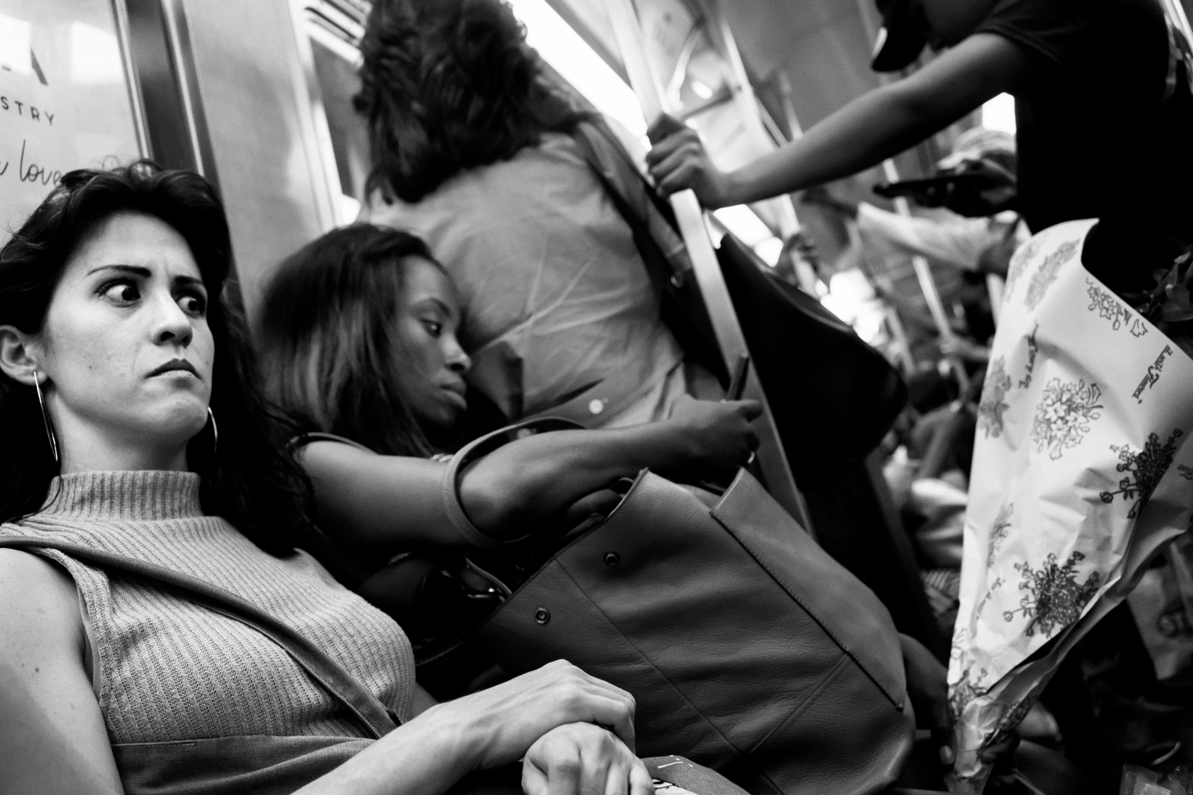 Black and white photo of passengers seated and standing inside a crowded train carriage, some holding bags and flowers; a woman seated in the foreground has an annoyed look on her face.