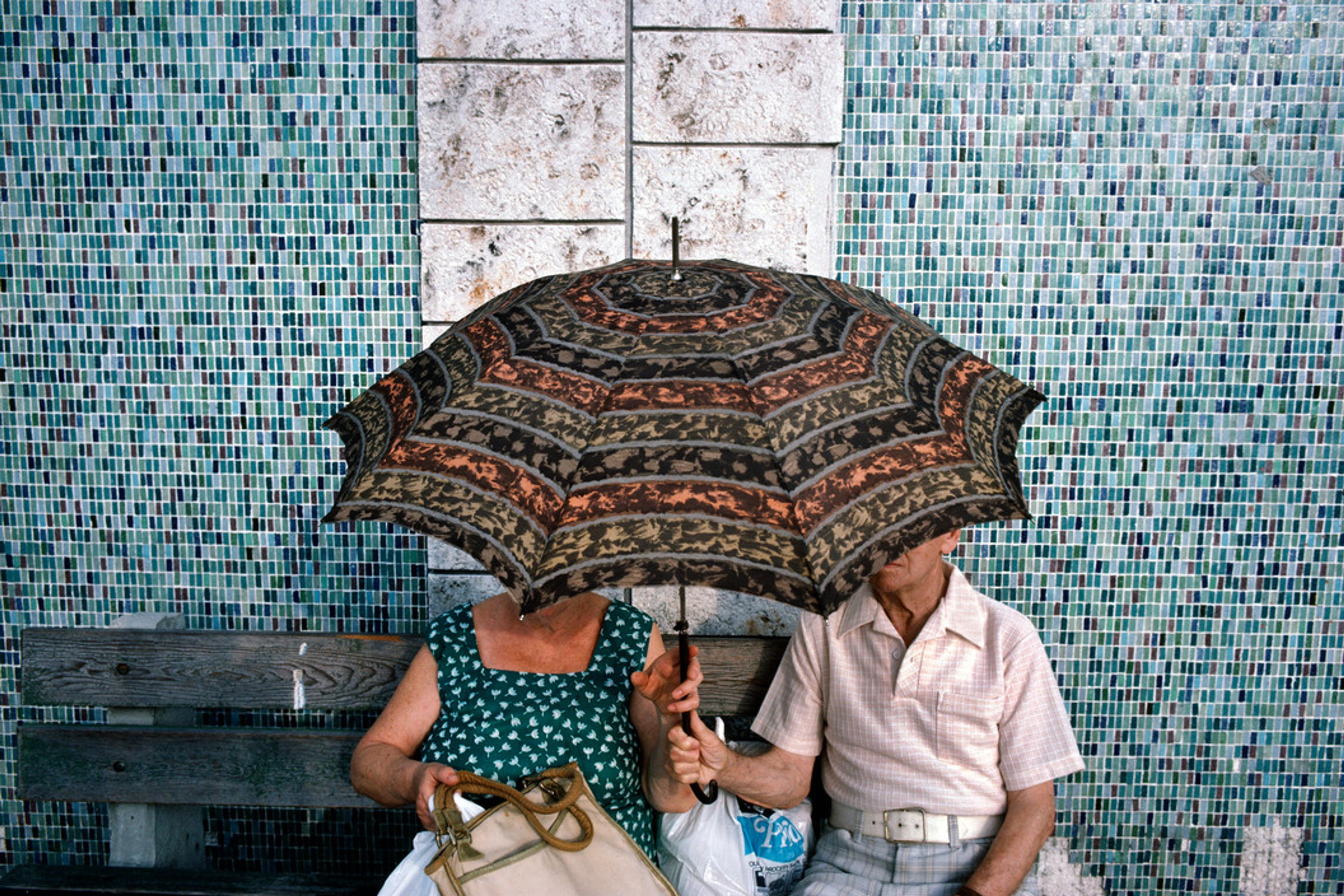 An elderly couple sitting on a bench under a patterned umbrella, with a mosaic-tiled wall in the background.