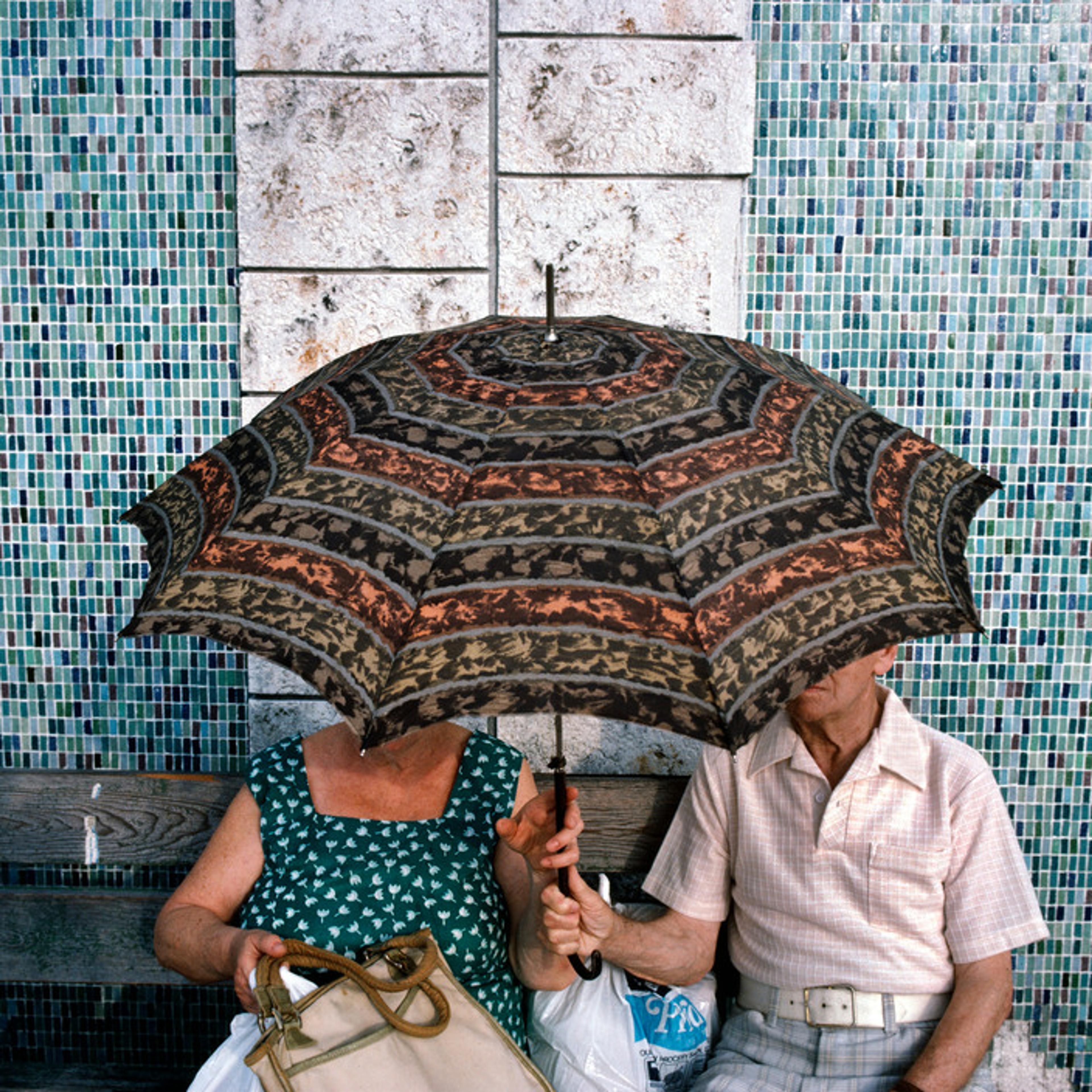 Una pareja de ancianos sentada en un banco bajo una sombrilla estampada, con una pared de mosaicos al fondo.