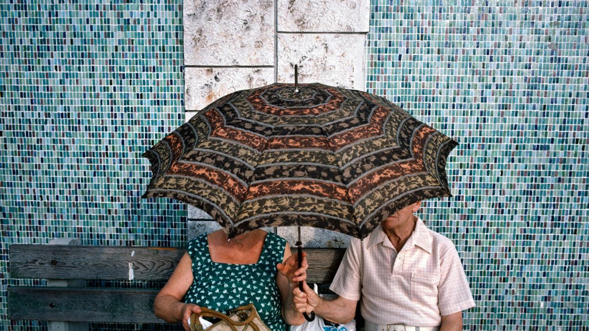 An elderly couple sitting on a bench under a patterned umbrella, with a mosaic-tiled wall in the background.