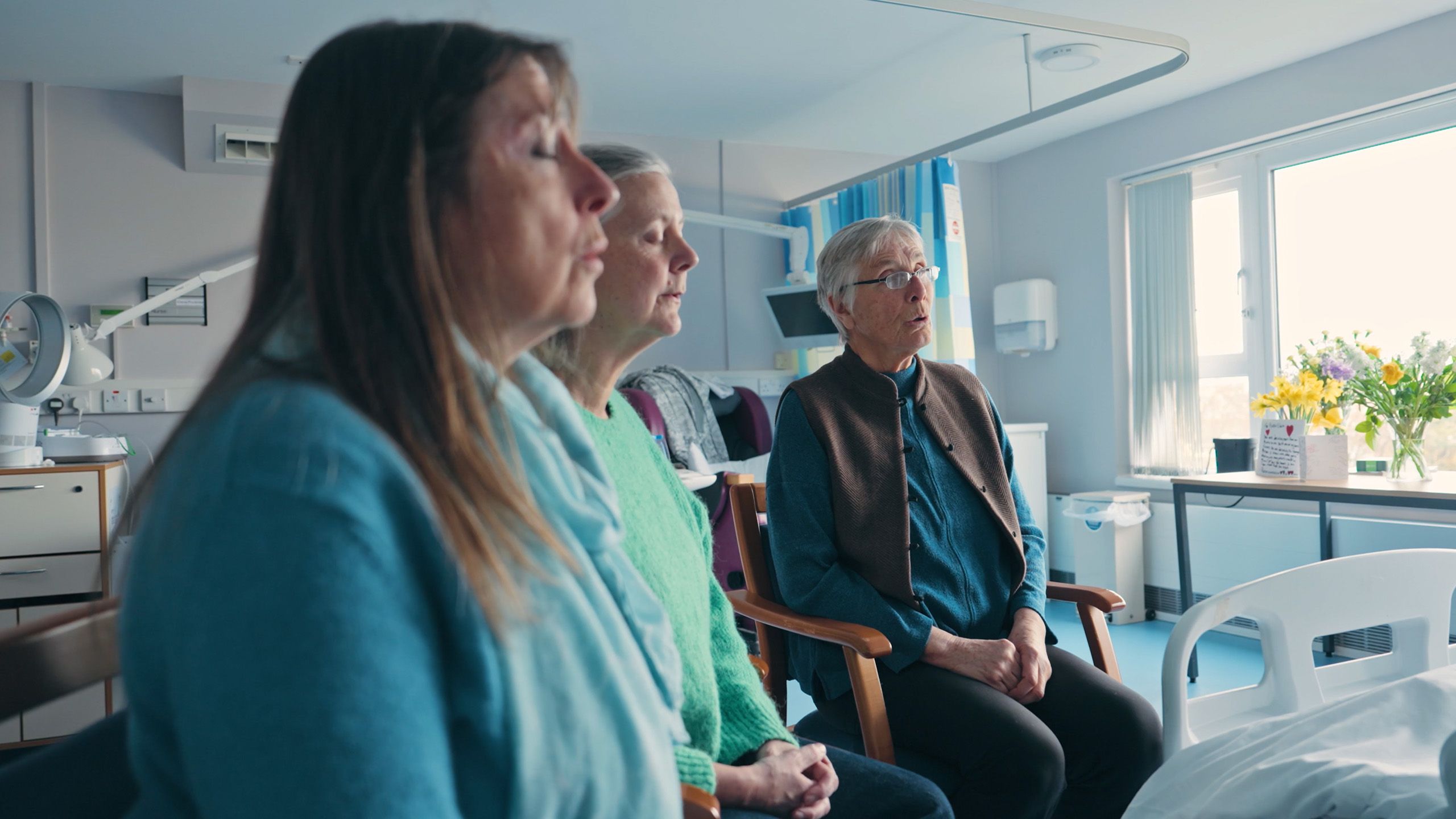 Three people sitting in a hospital room, singing with their eyes closed, with flowers on the windowsill in the background.