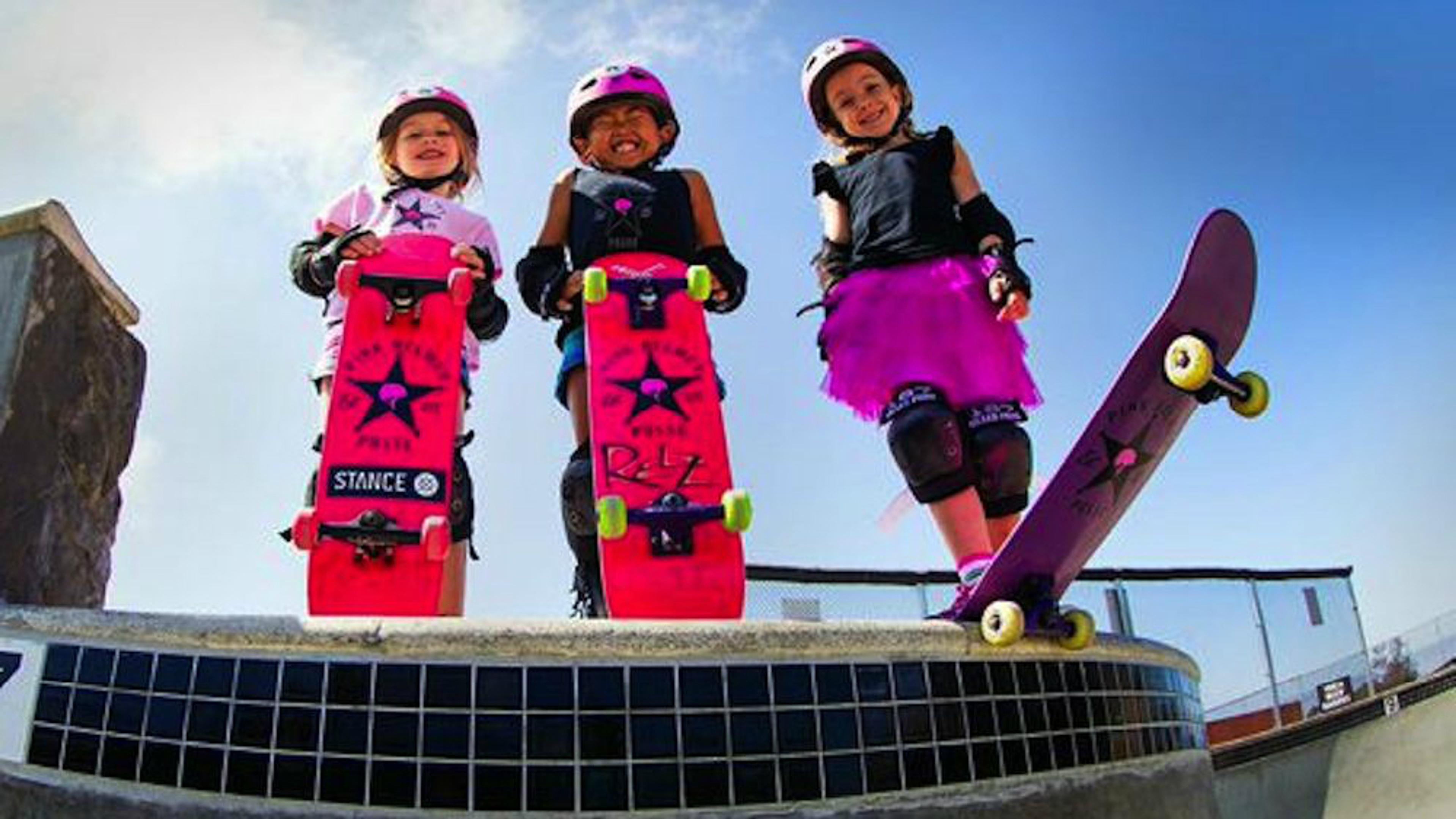 Three girls with pink skateboards at a skate park, smiling and wearing helmets, standing on a ledge ready to skateboard.
