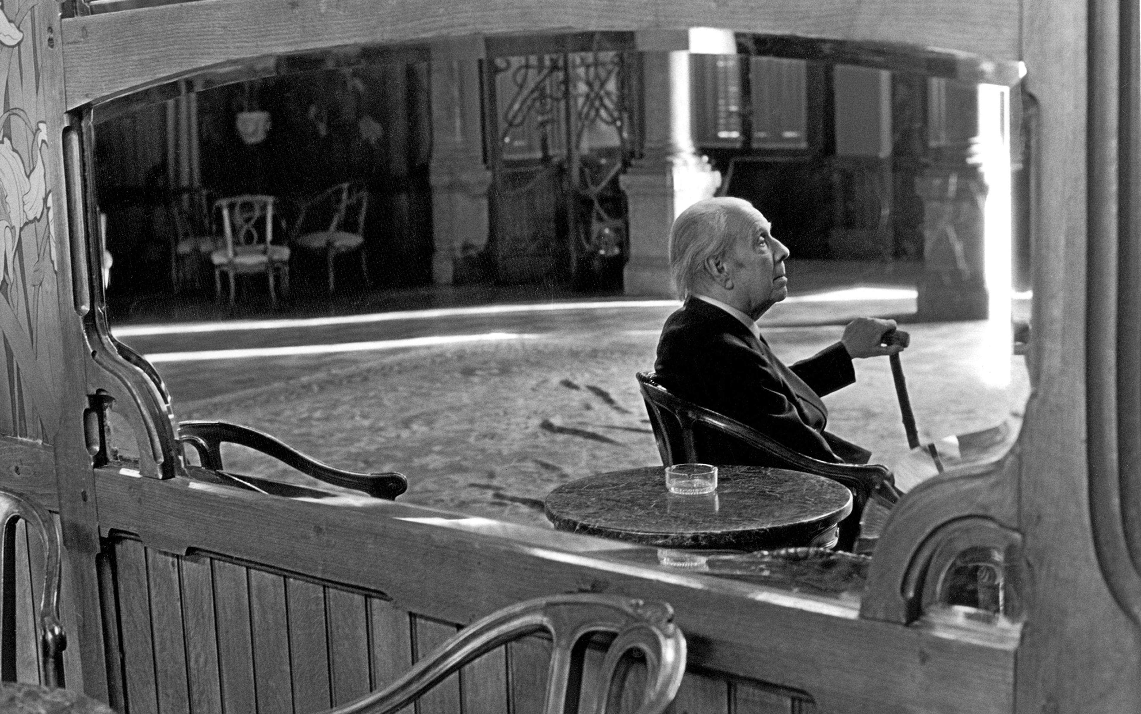 Elderly man sitting at a table in a spacious, elegant room, holding a cane and looking contemplatively to the right.