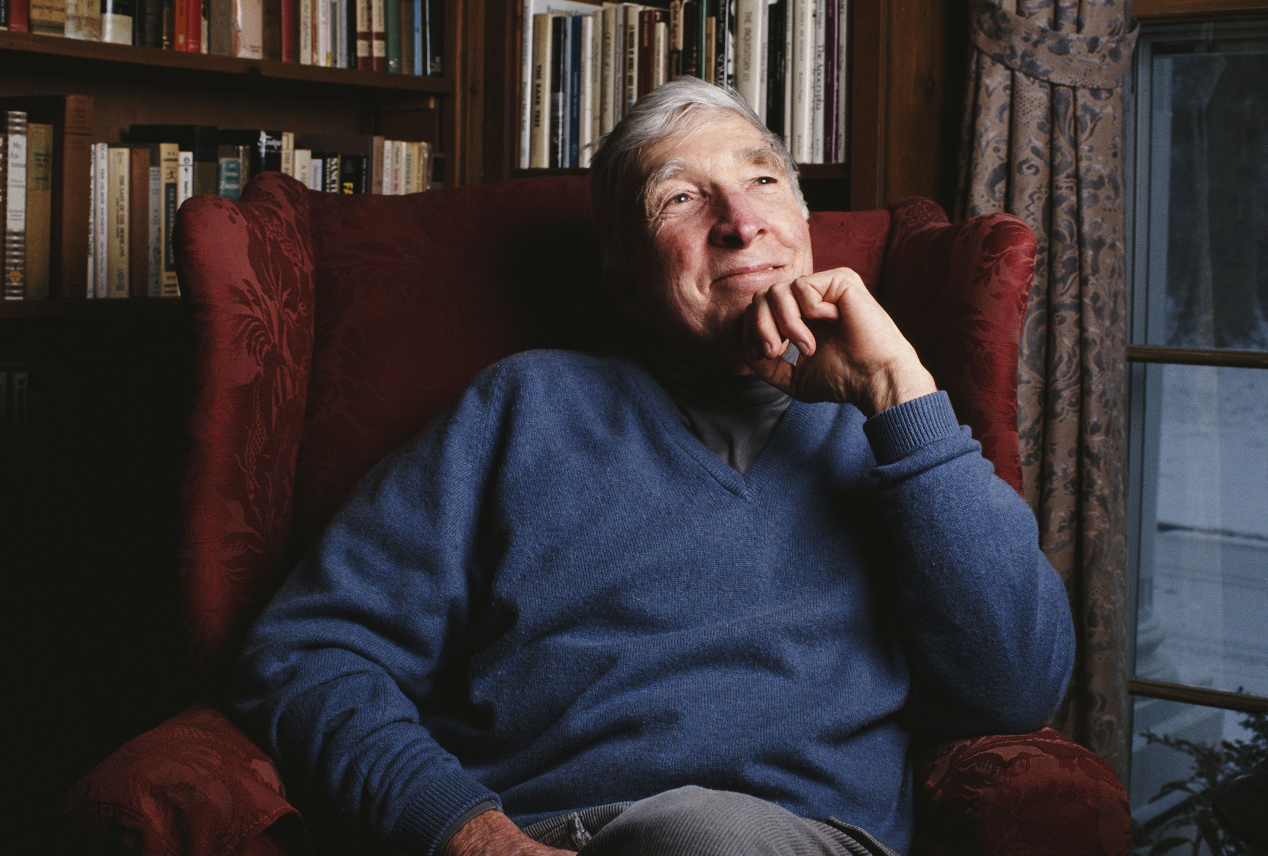 An elderly man in a blue jumper seated in a red armchair, surrounded by bookshelves and a window with curtains.