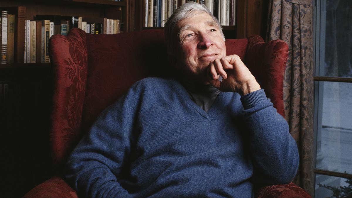 An elderly man in a blue jumper seated in a red armchair, surrounded by bookshelves and a window with curtains.